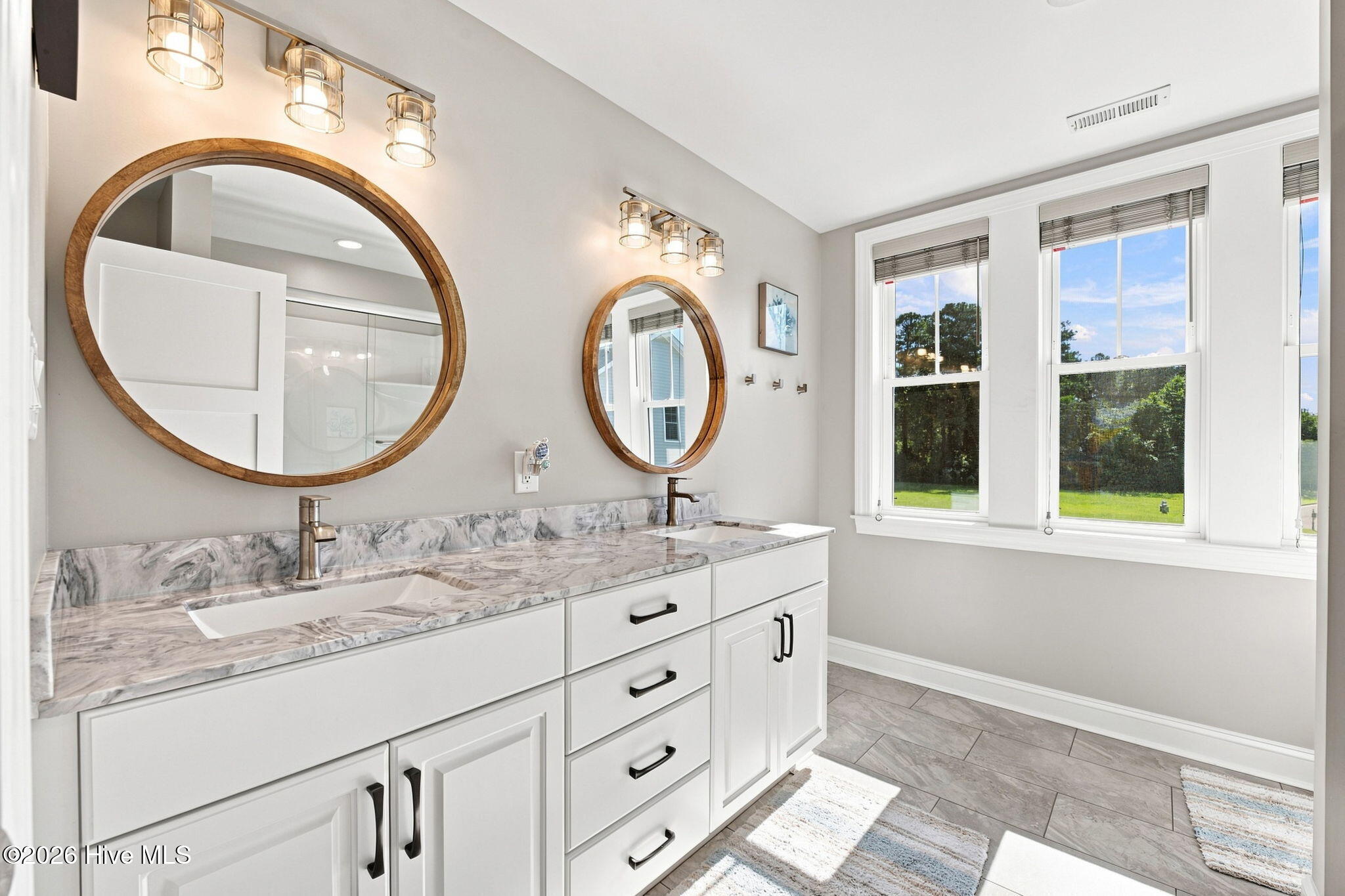 Bathroom with a marble double vanity, round wood-framed mirrors, and large windows overlooking a lawn.