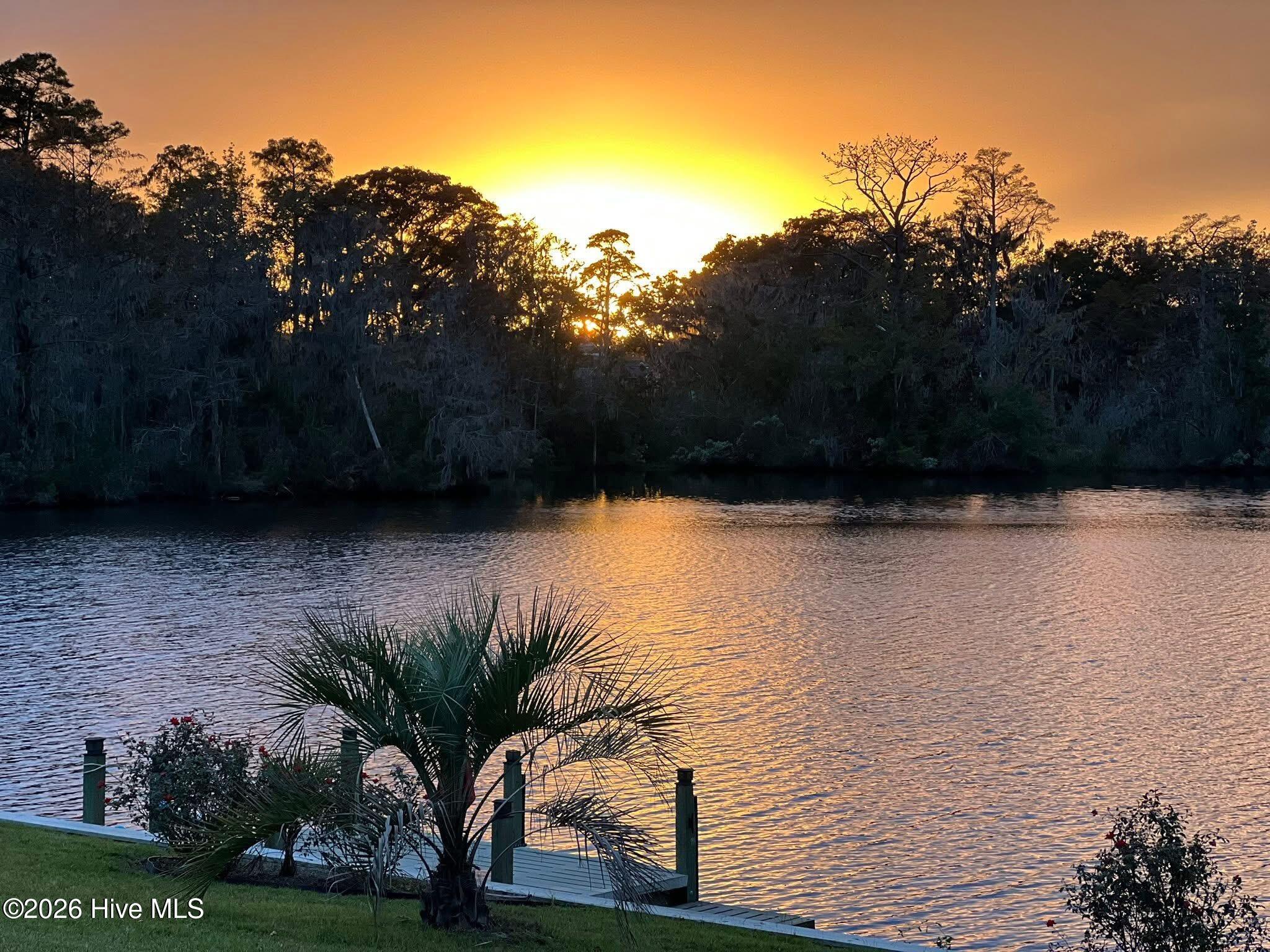 Waterfront at sunset, with a small dock, palm-like landscaping, and golden reflections on the water.