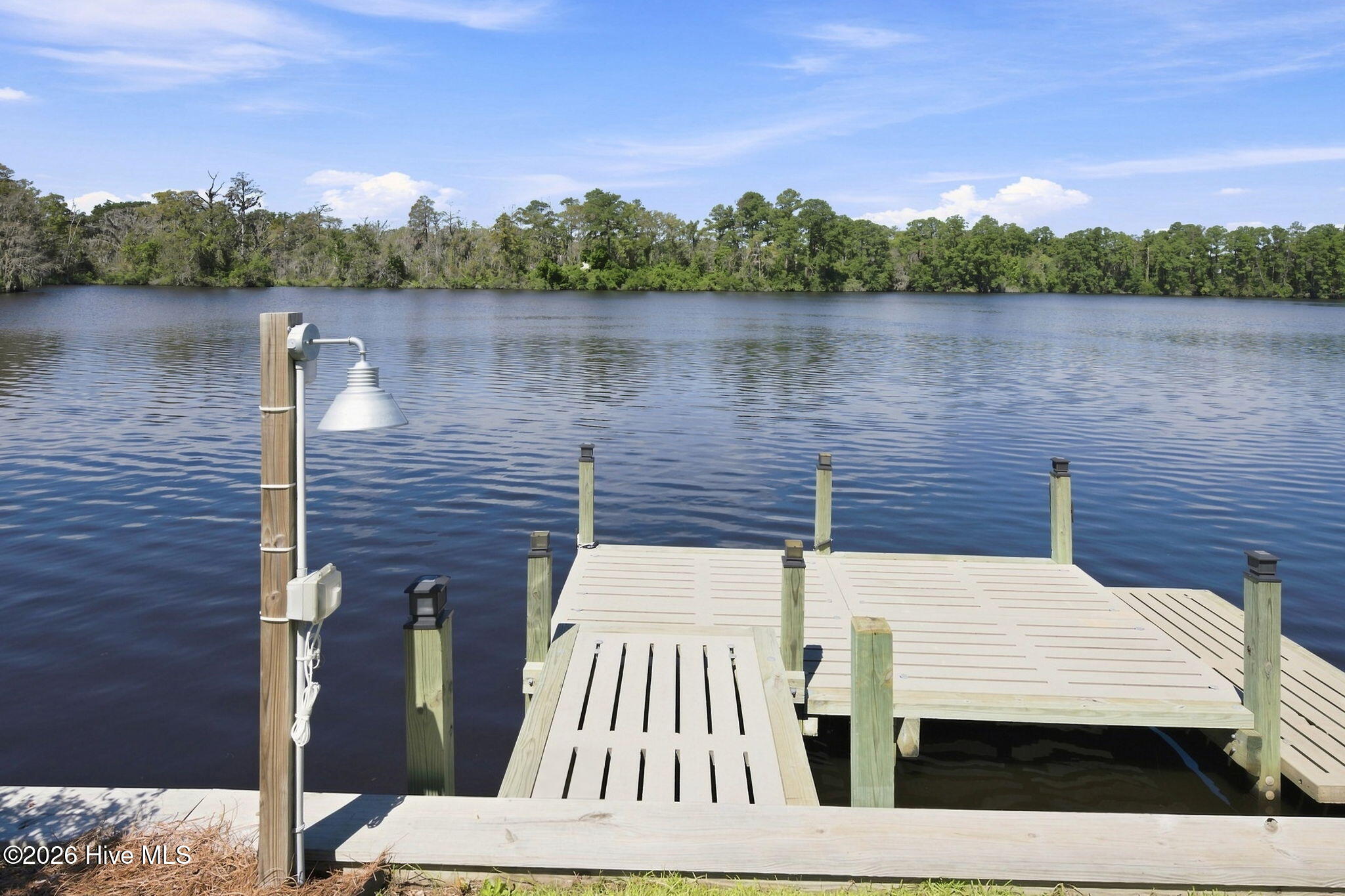 Dock extending into a calm lake, with a lamp and wooded shoreline across the water.