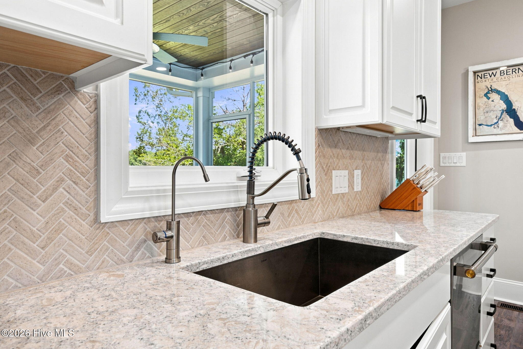 Kitchen sink with two faucets, pale stone counters, herringbone backsplash, and a large window overlooking trees.