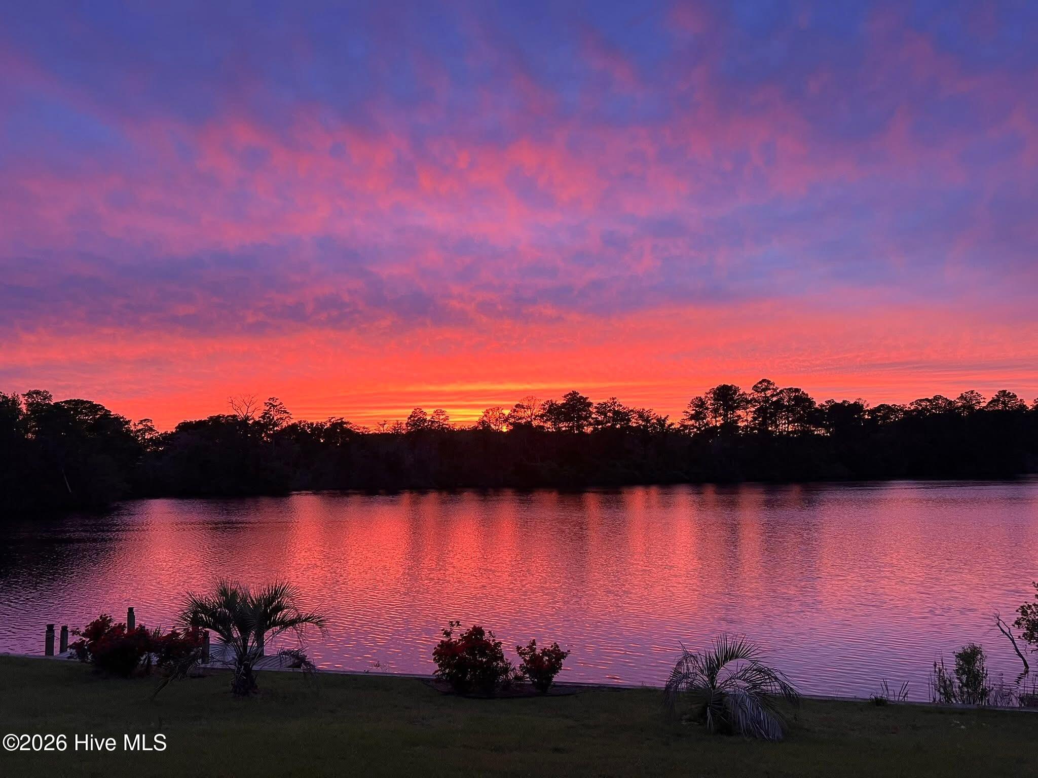 Sunset colors reflect on a waterfront, with a tree-lined shore and landscaped lawn.