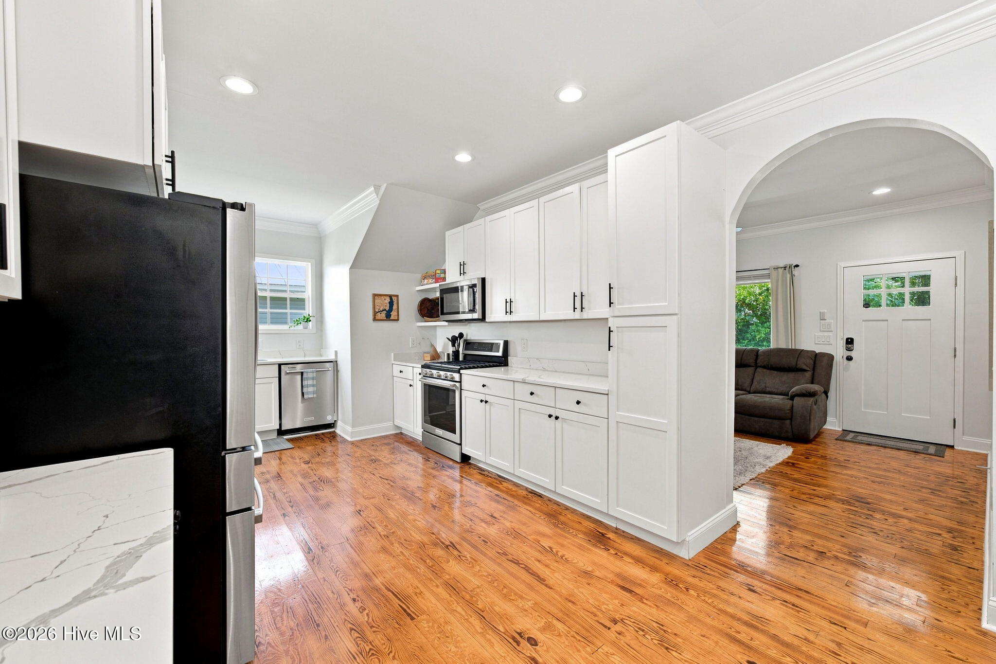 Kitchen with white cabinets, stainless steel appliances, warm wood floors, and an arched opening to the living area.