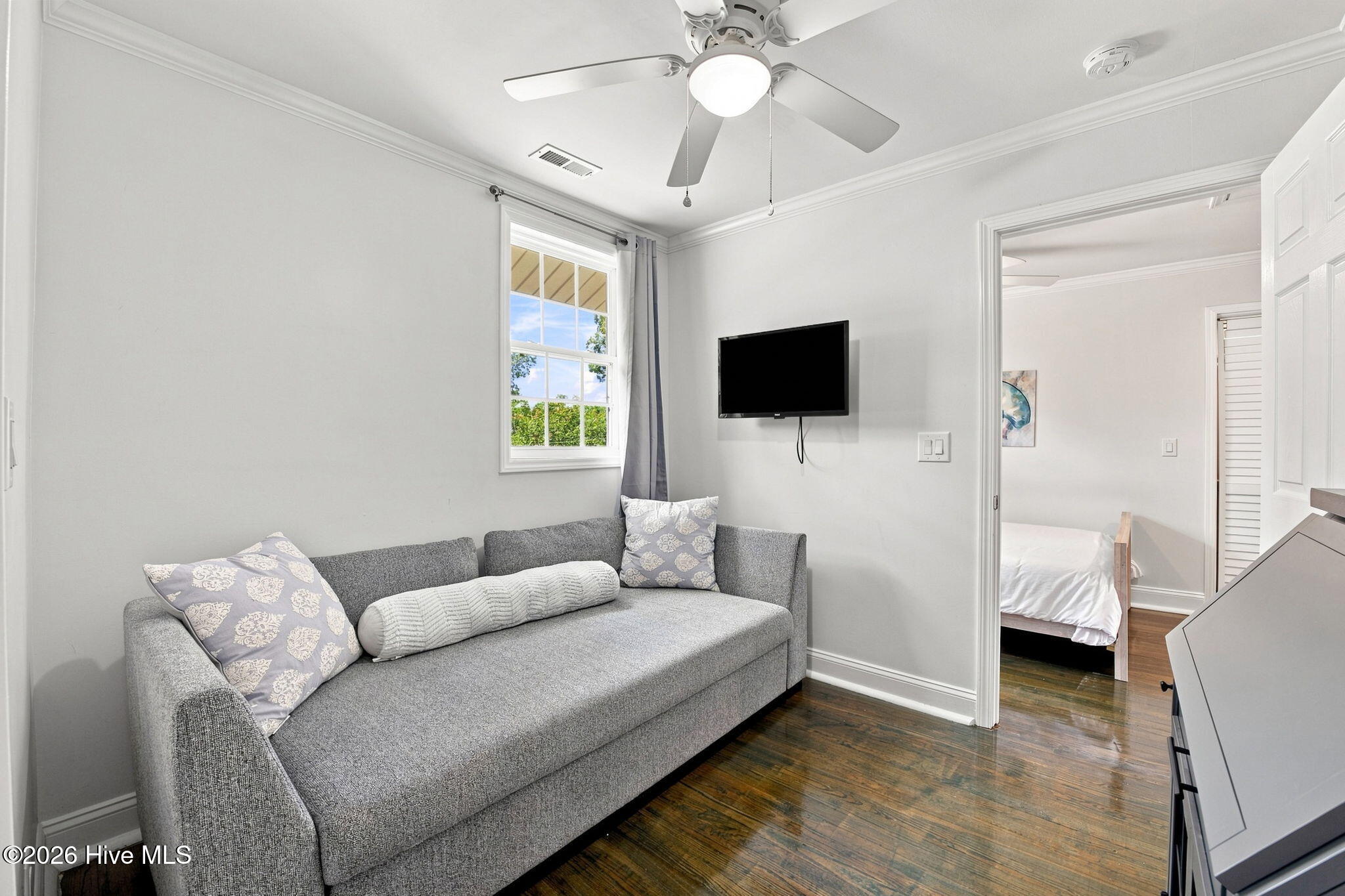 Furnished sitting room with a gray sofa, wall-mounted television, and doorway to an adjoining bedroom.
