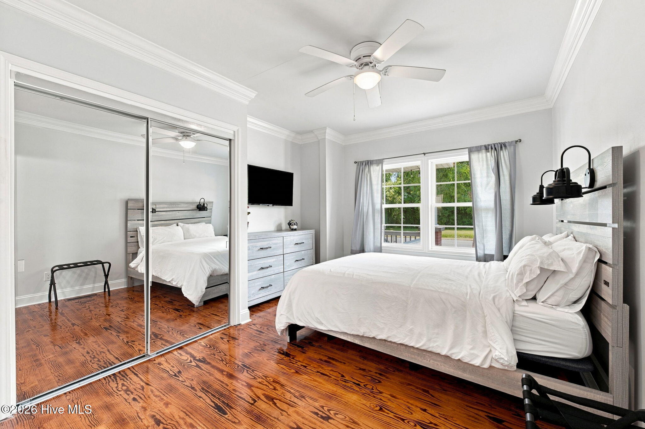Bedroom with a white bed, mirrored closet doors, and polished reddish hardwood floors.