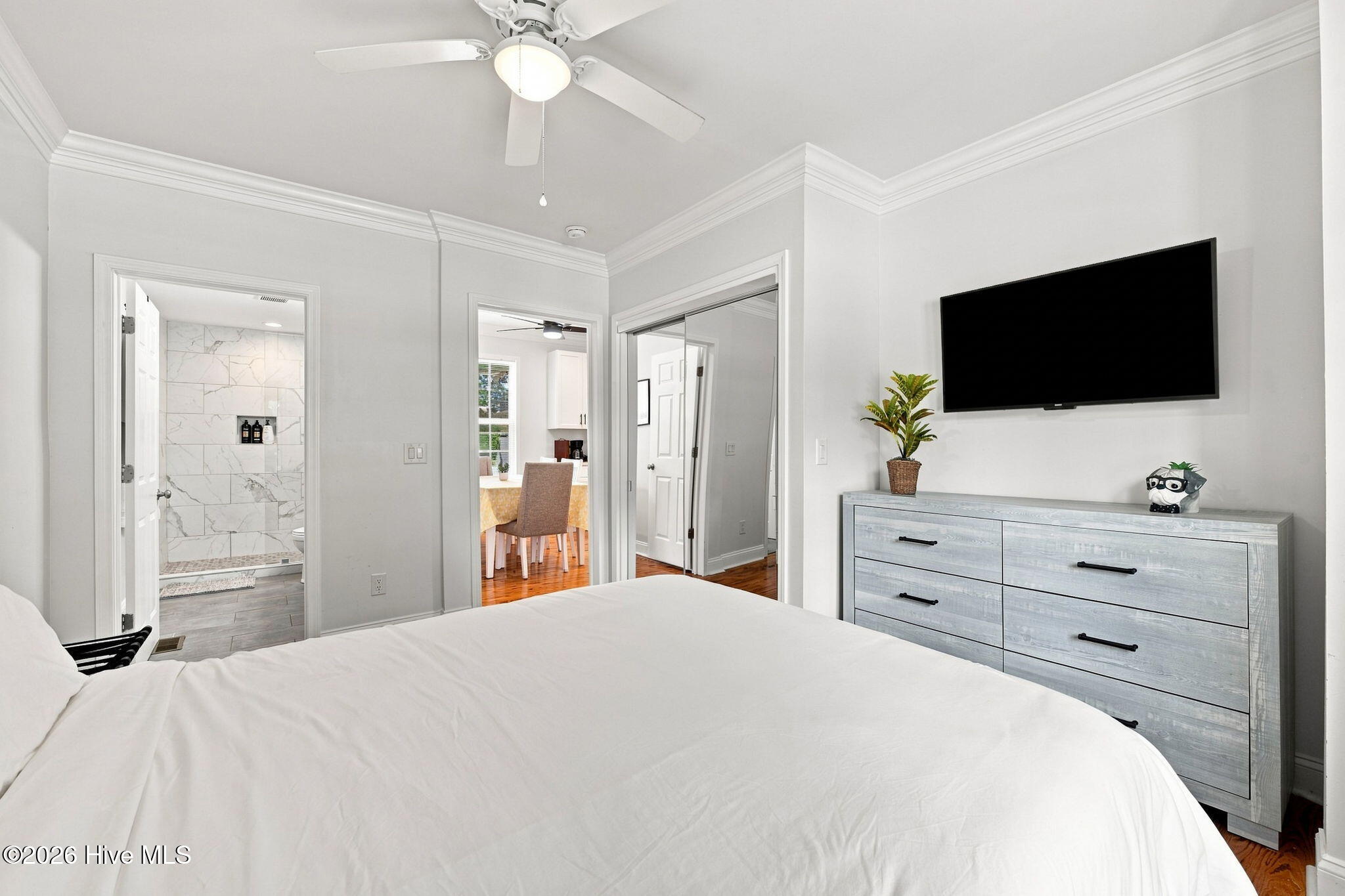 Bedroom with a white bed, gray dresser, wall-mounted television, and open doorway to a tiled shower.