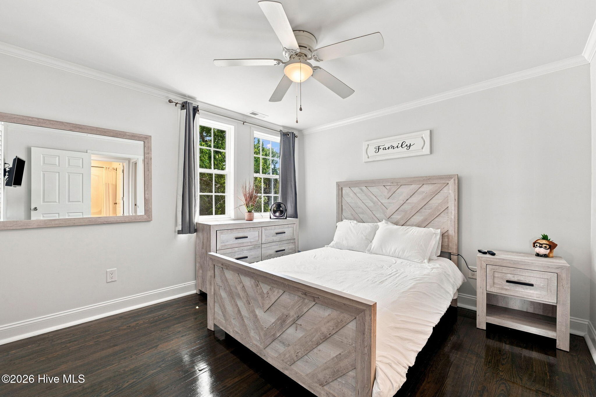 Bedroom with a white bed, geometric pale wood furniture, dark floors, and two sunny windows.