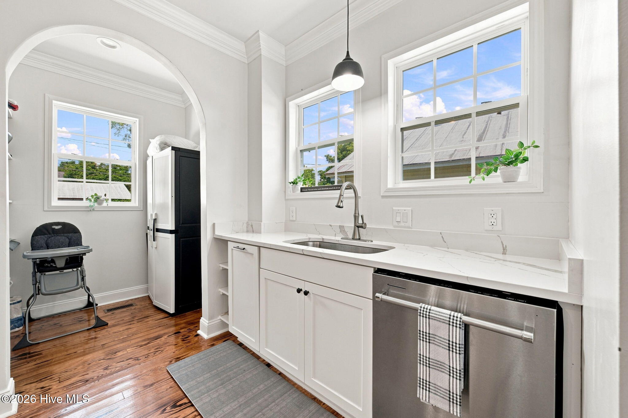 Bright white kitchen with marble-look counters, stainless-steel dishwasher, large windows, and warm wood floors.
