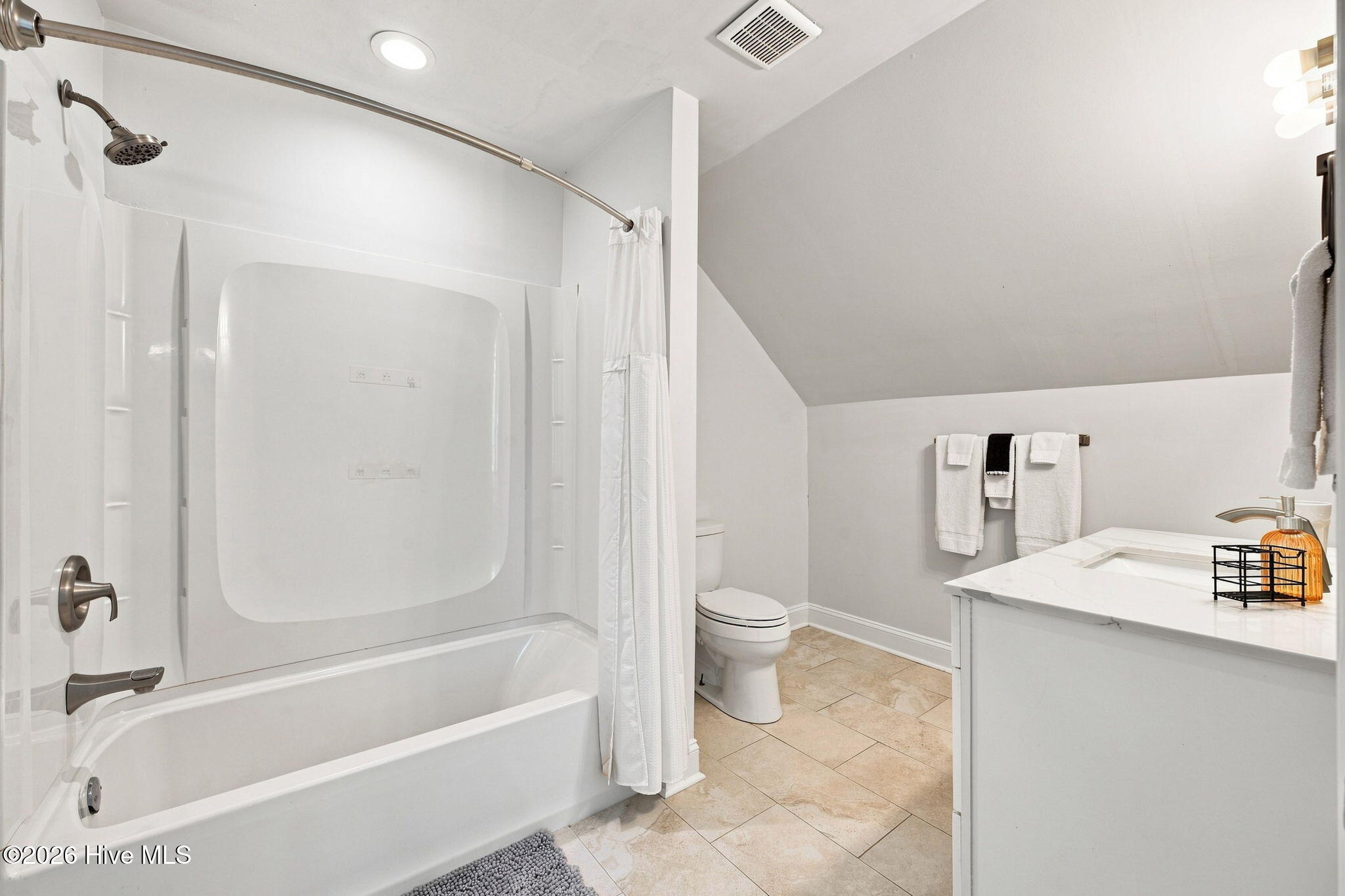 Bathroom with a white tub and shower, tiled floor, and vanity beneath a sloped ceiling.