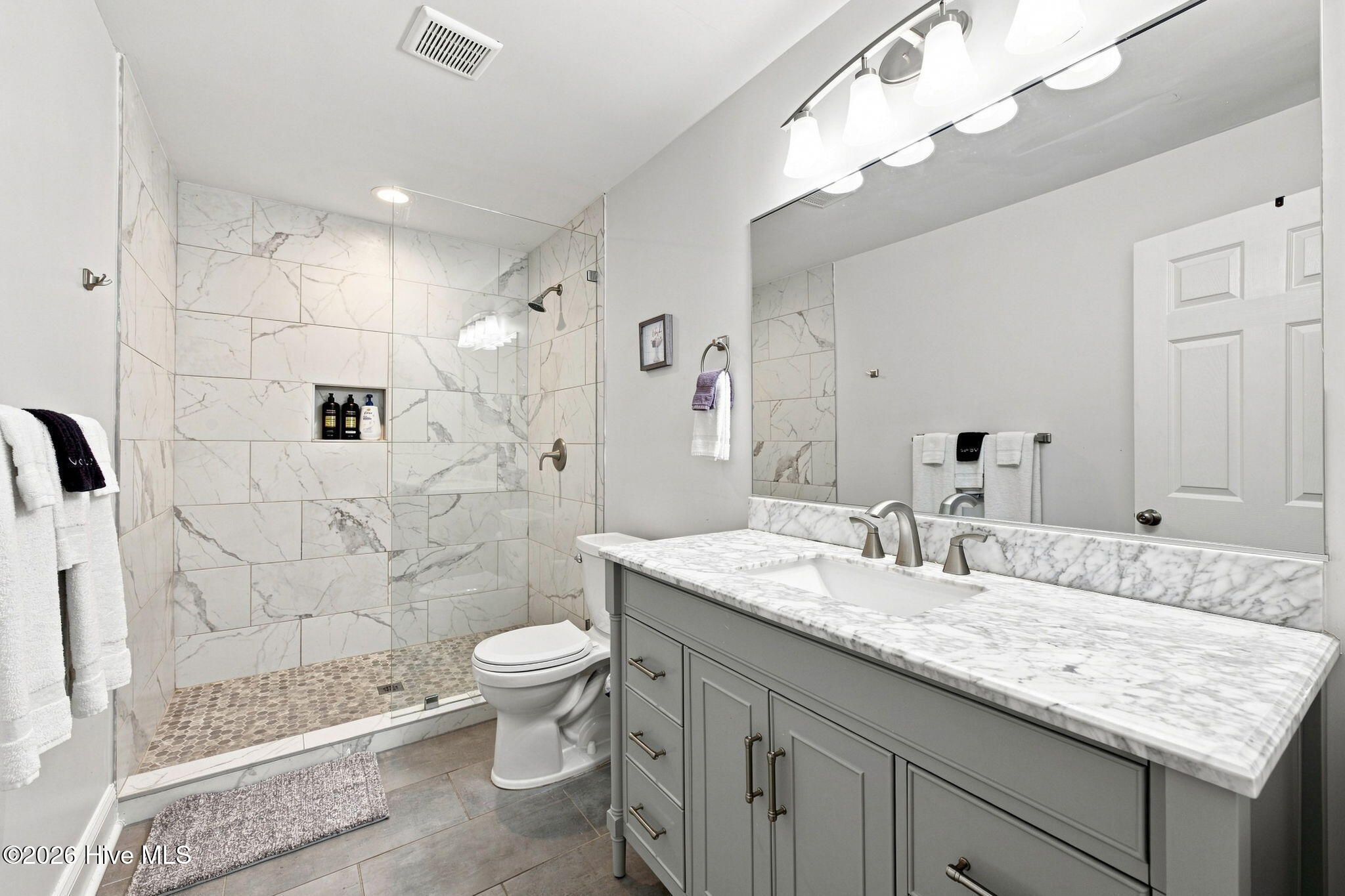 Bathroom with a marble-look tiled shower, pebble floor, and gray vanity with a white countertop.