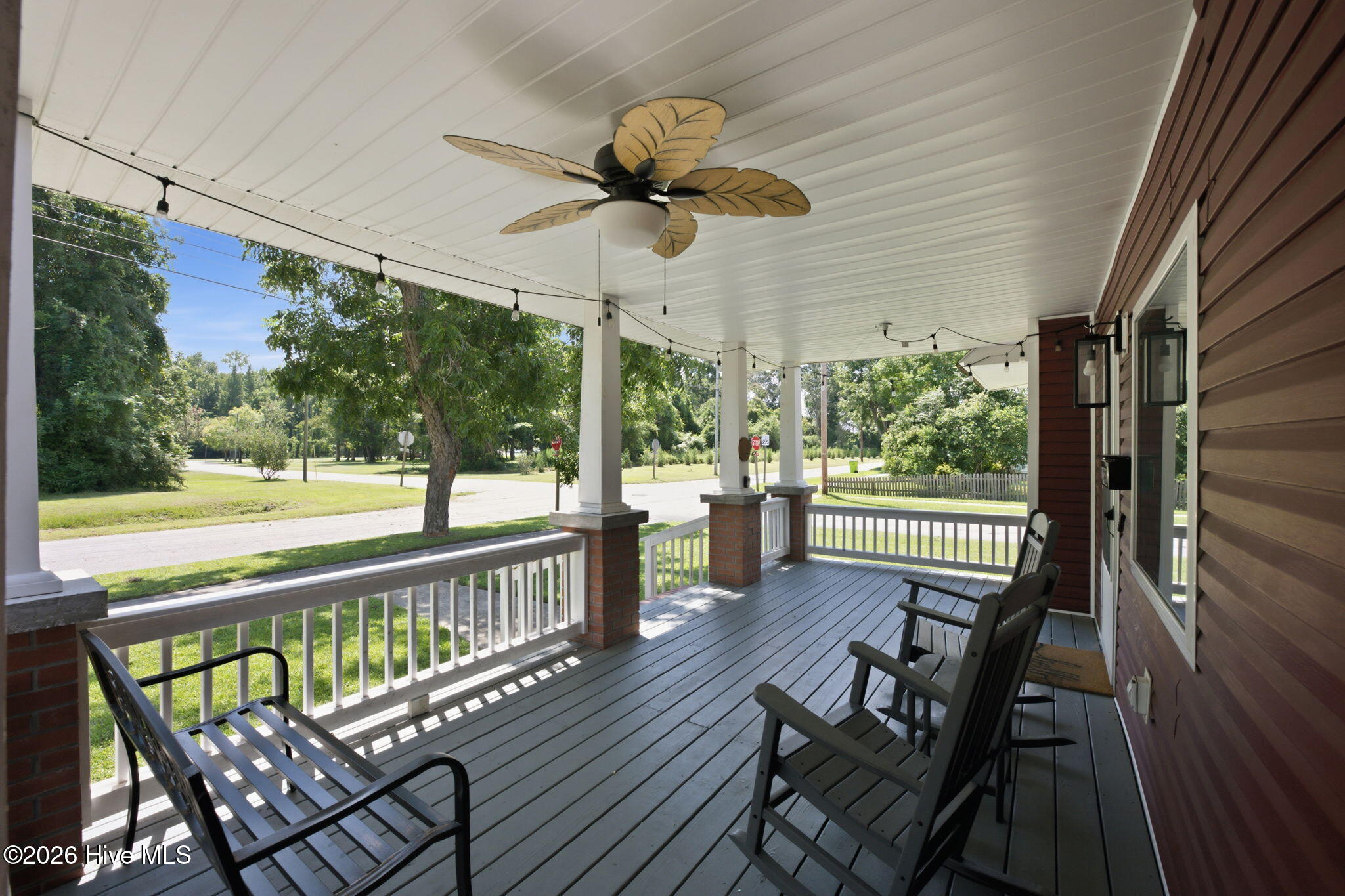 Covered front porch with rocking chairs, a leaf-shaped ceiling fan, and a view of the tree-lined street.