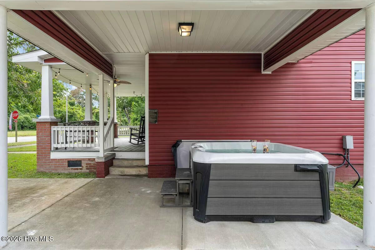 Covered patio with a large hot tub beside a red-sided home and white-railed porch.