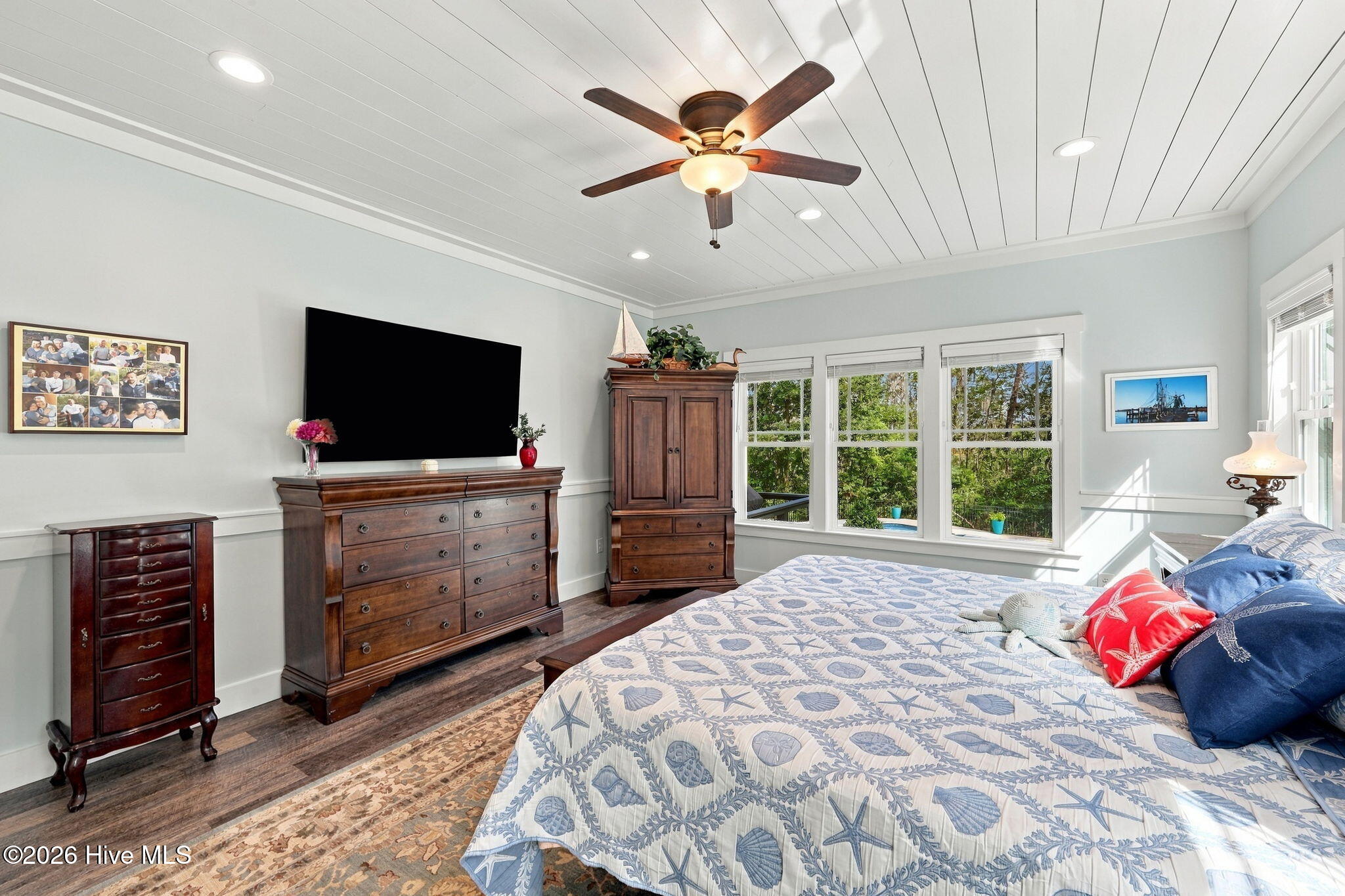 Bedroom with seashell-patterned bedding, dark wood dressers, and windows overlooking trees.