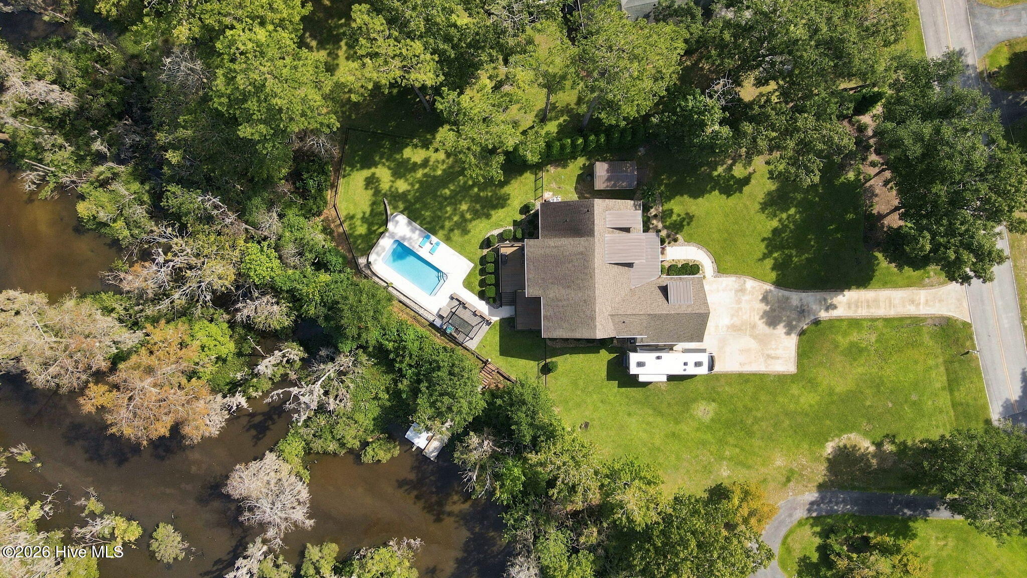Aerial view of a creekside home with a fenced backyard, swimming pool, shed, and long driveway.