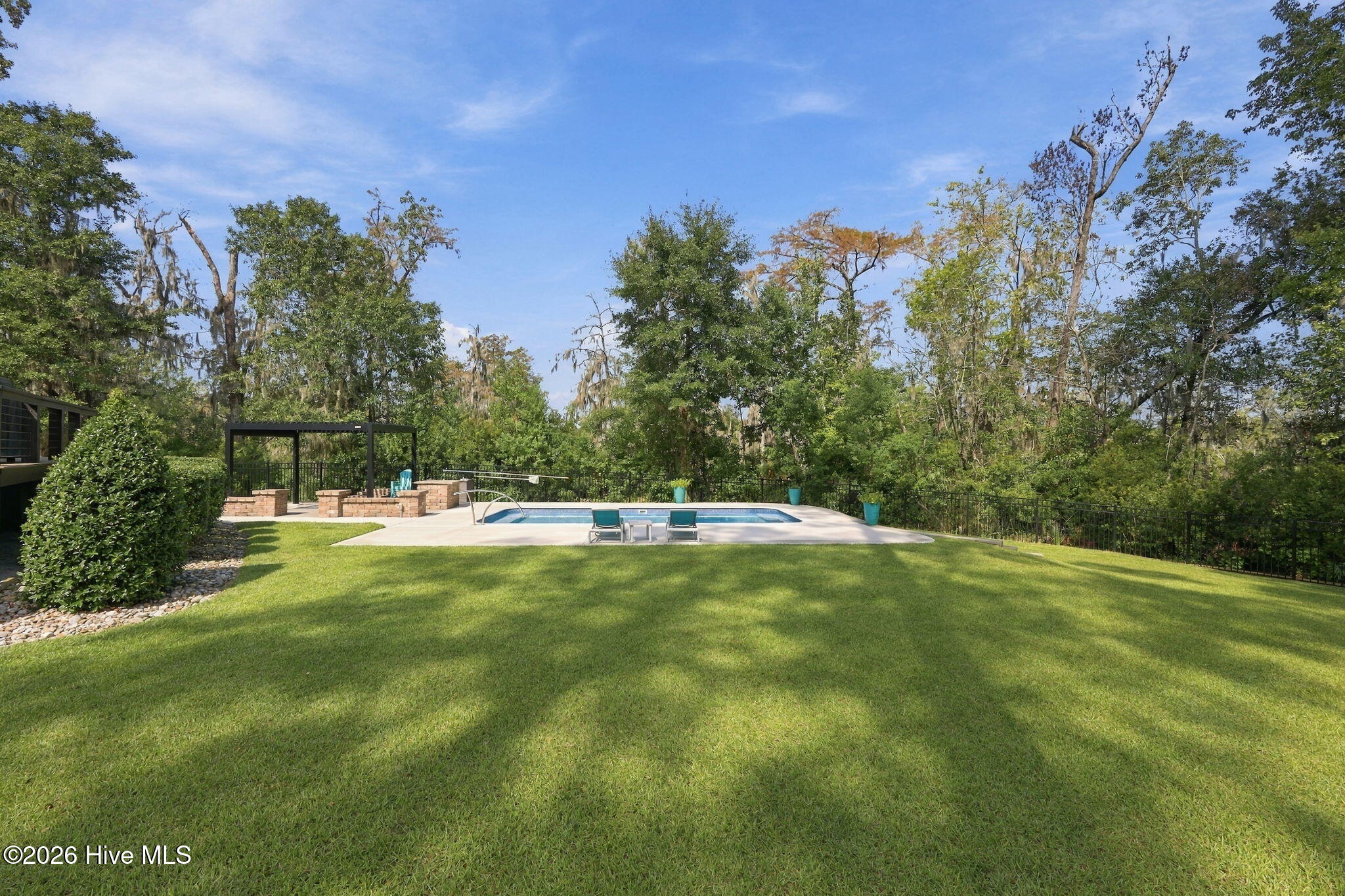 Backyard swimming pool beside a broad lawn, with poolside chairs and a pergola near mature trees.