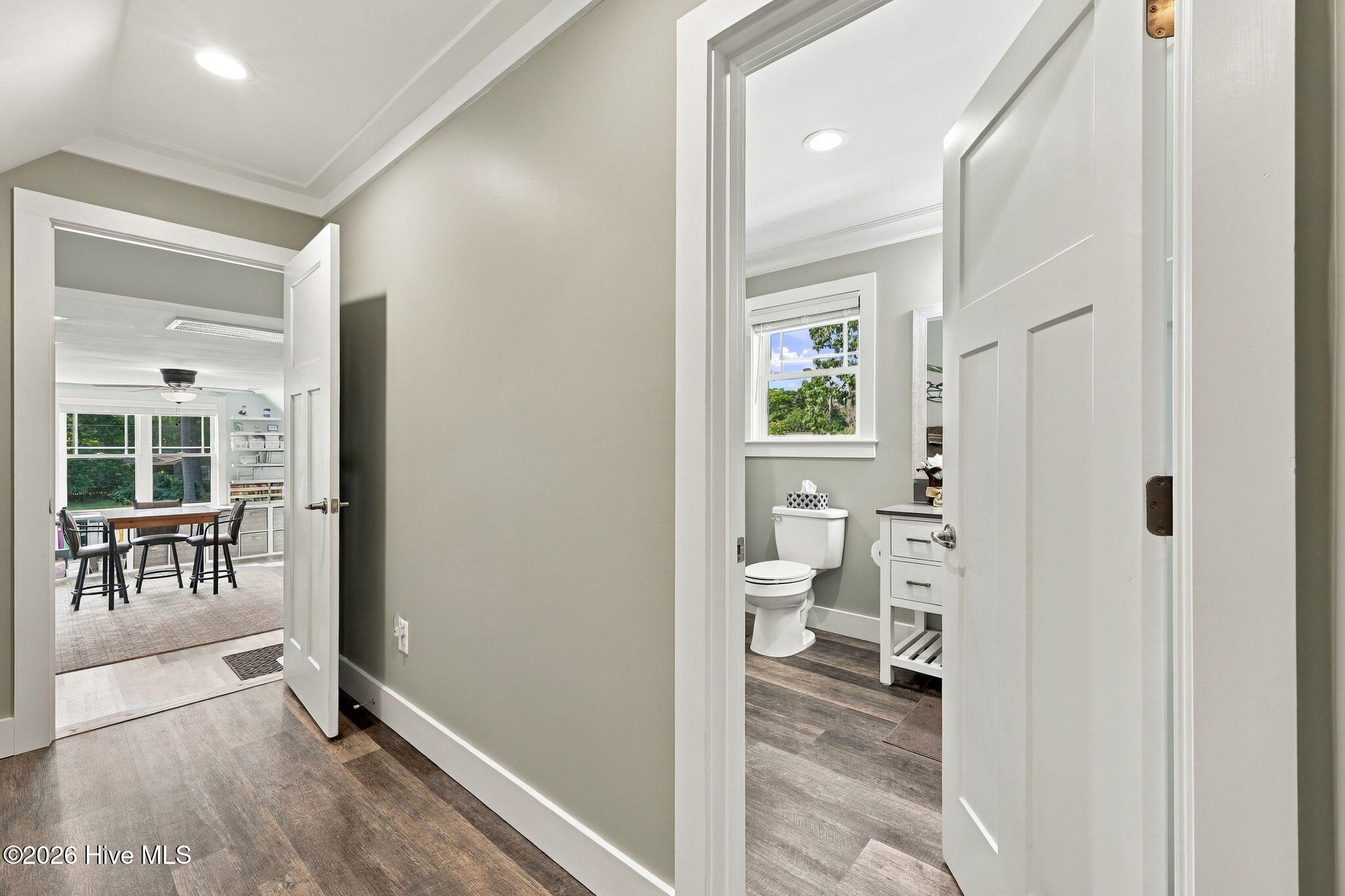 Hallway with open doors to a bathroom and a bright dining area with large windows.