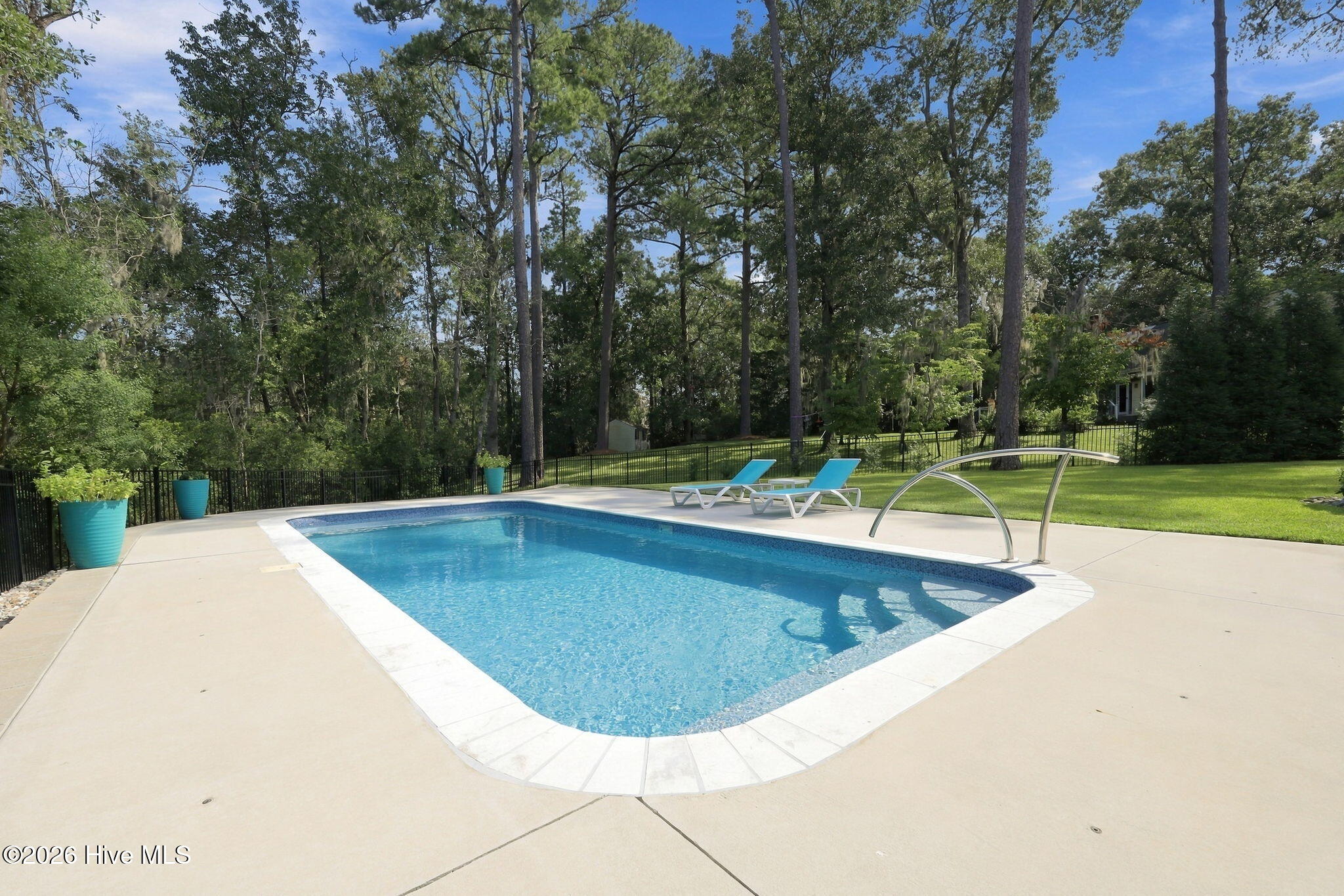 Backyard swimming pool with blue lounge chairs, a curved metal handrail, and mature trees.