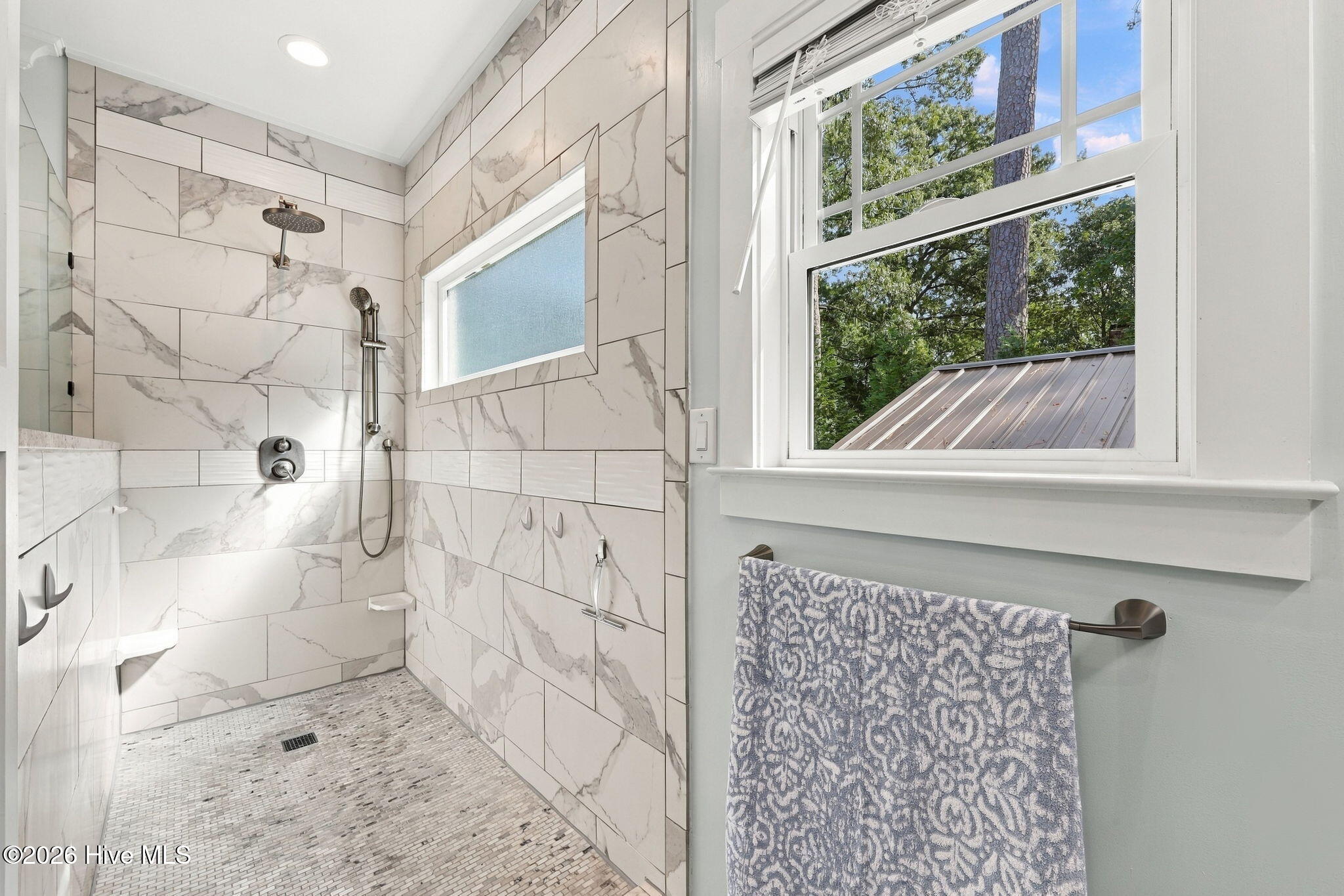 Bathroom with a marble-look tiled walk-in shower and large window overlooking leafy trees.