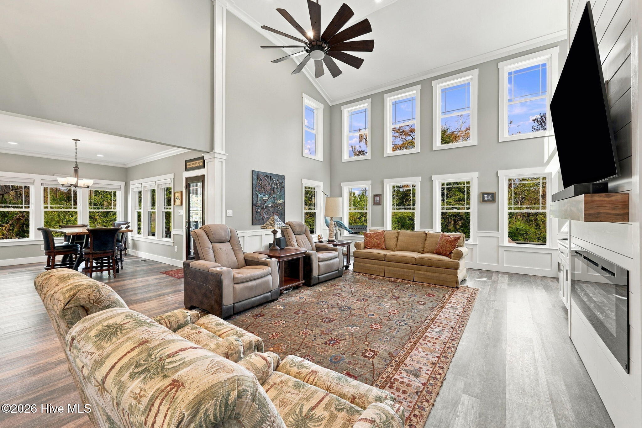 Furnished living room with vaulted ceiling, tall windows, patterned rug, and wall-mounted television.