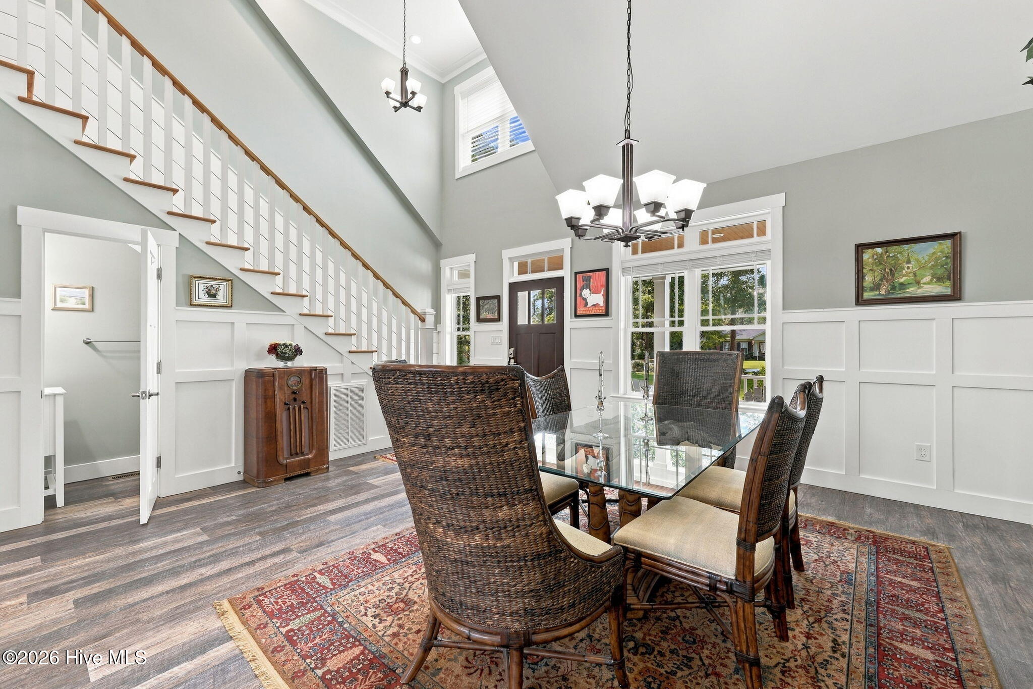 Dining room with a glass-top table, patterned rug, and staircase rising beneath a high ceiling.
