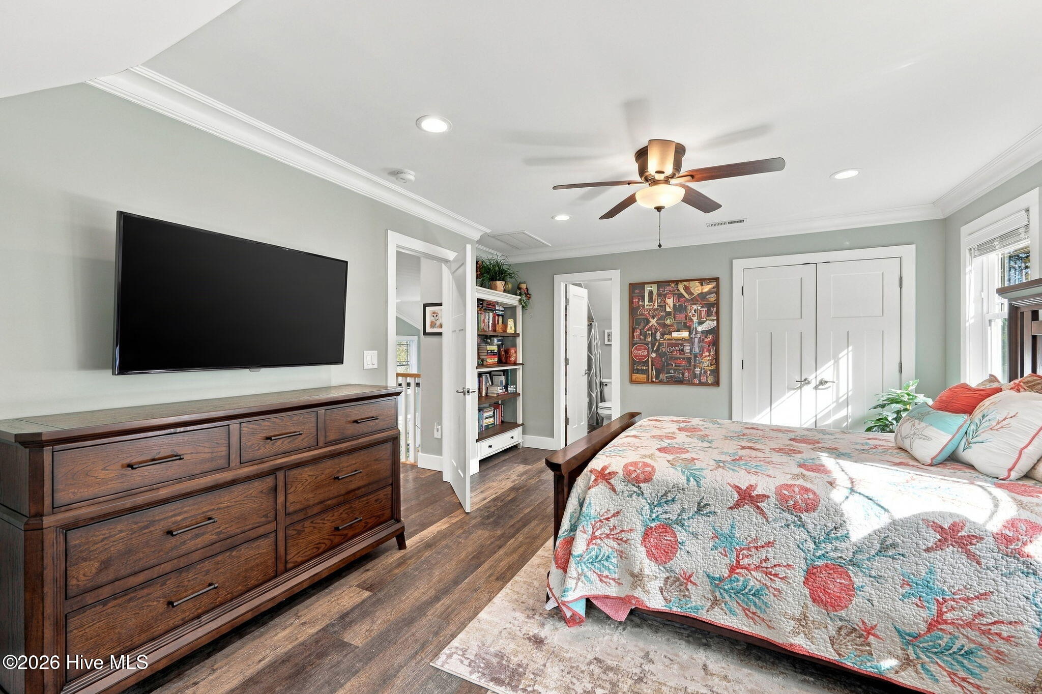 Furnished bedroom with a floral quilt, dark wood dresser, wall-mounted television, and bright window.