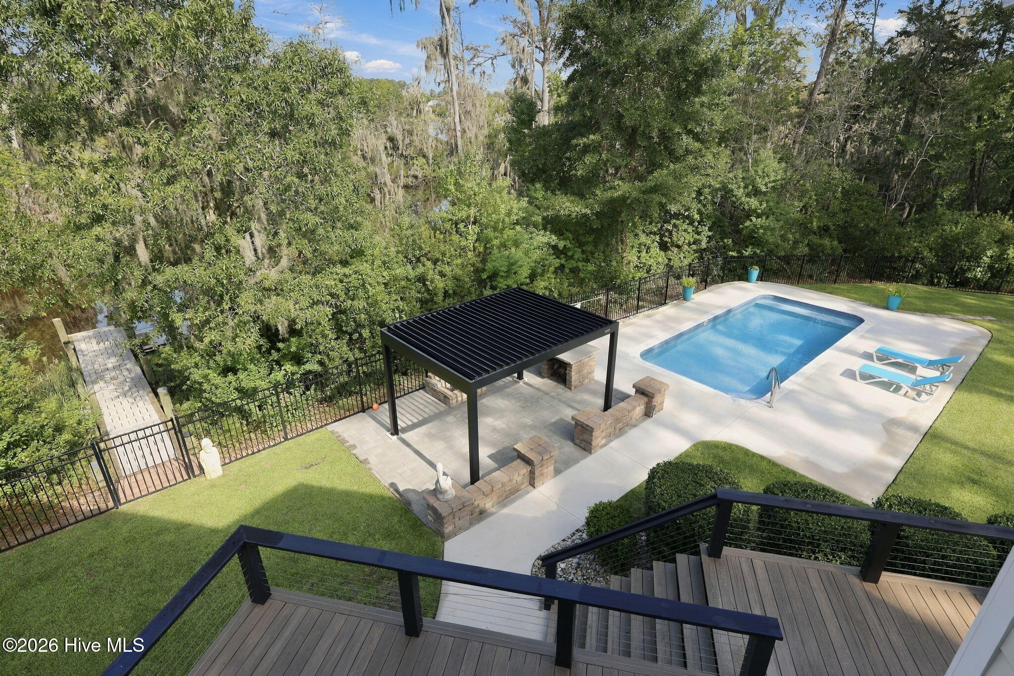 Backyard pool and paved patio with a black pergola, surrounded by trees.