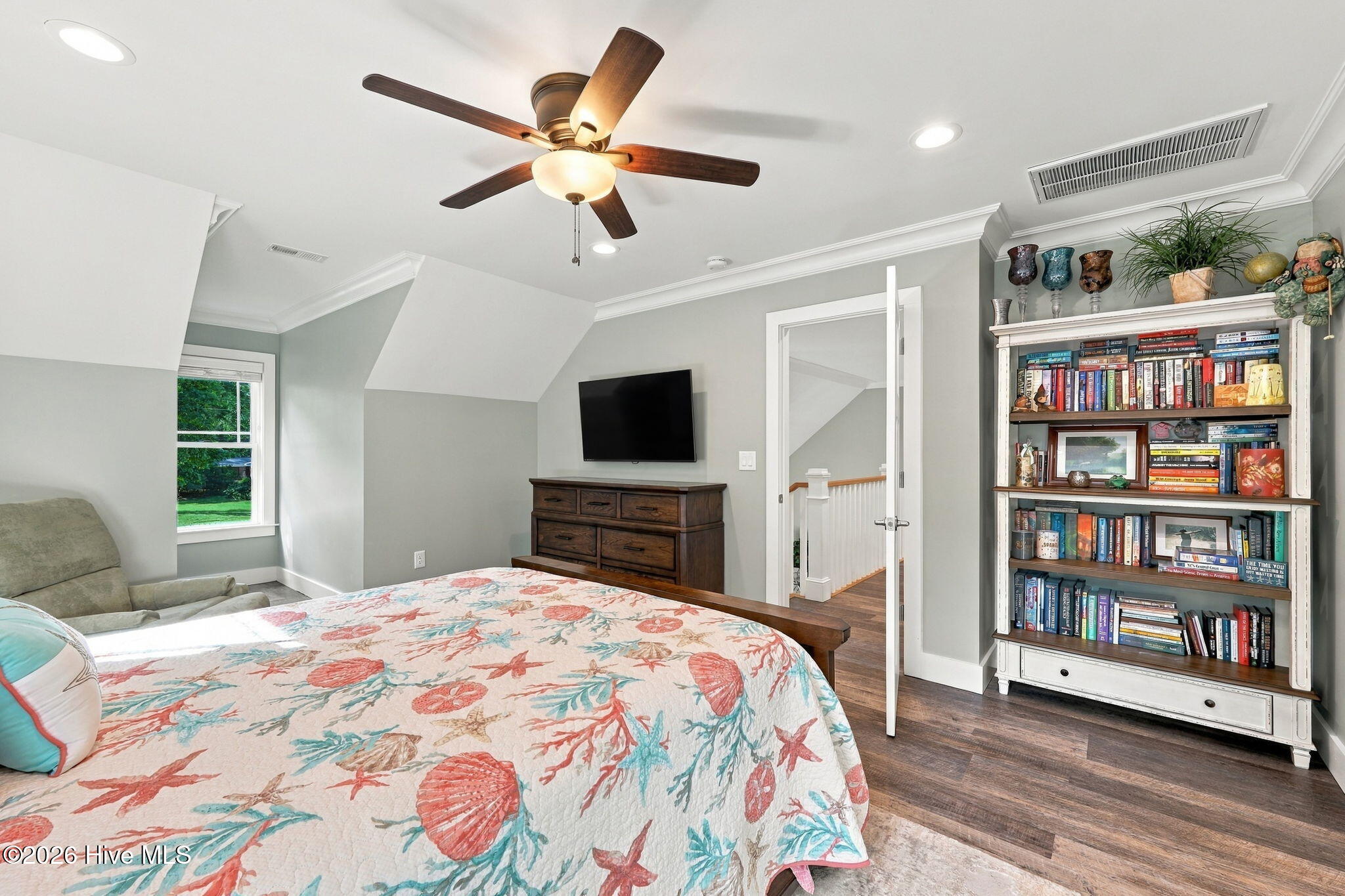 Bedroom with a colorful coastal-patterned quilt, ceiling fan, television, and book-filled bookcase.