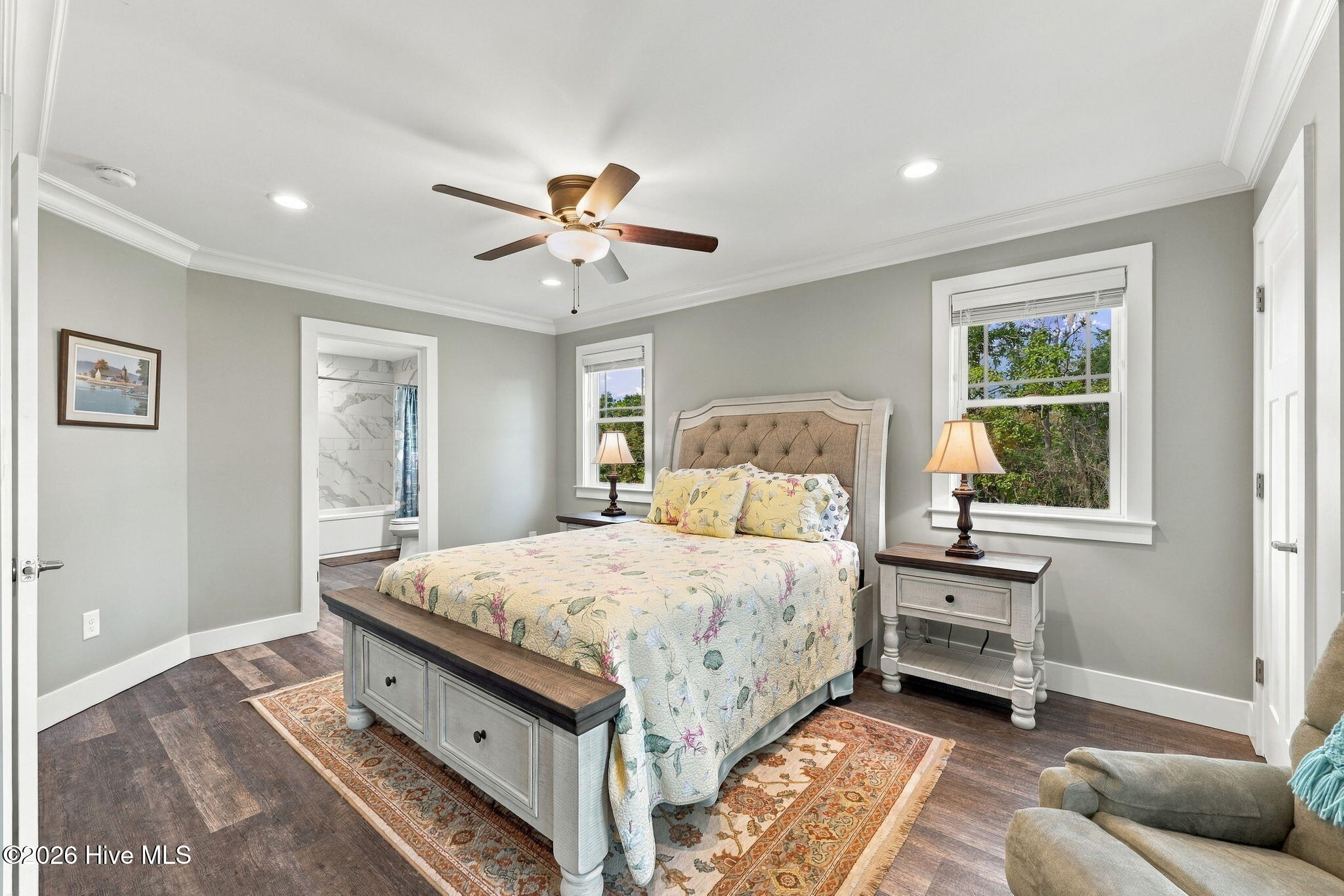 Bedroom with a floral-covered bed, tufted headboard, dark wood floors, and windows overlooking trees.
