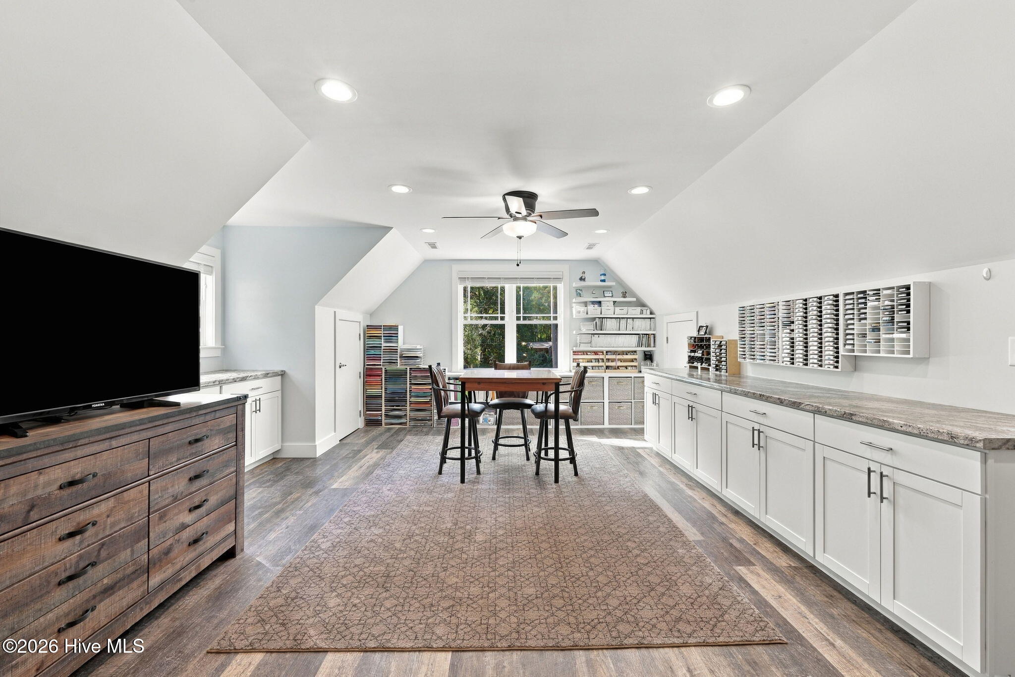 Attic craft room with a central worktable, extensive wall storage, white cabinets, and a window overlooking trees.