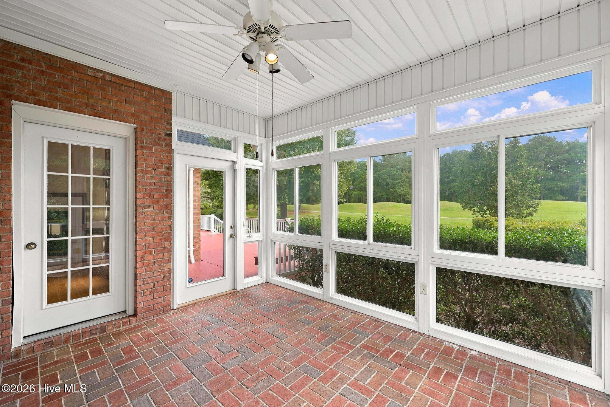 Sunroom with brick flooring, a ceiling fan, and large windows overlooking a green lawn and trees.