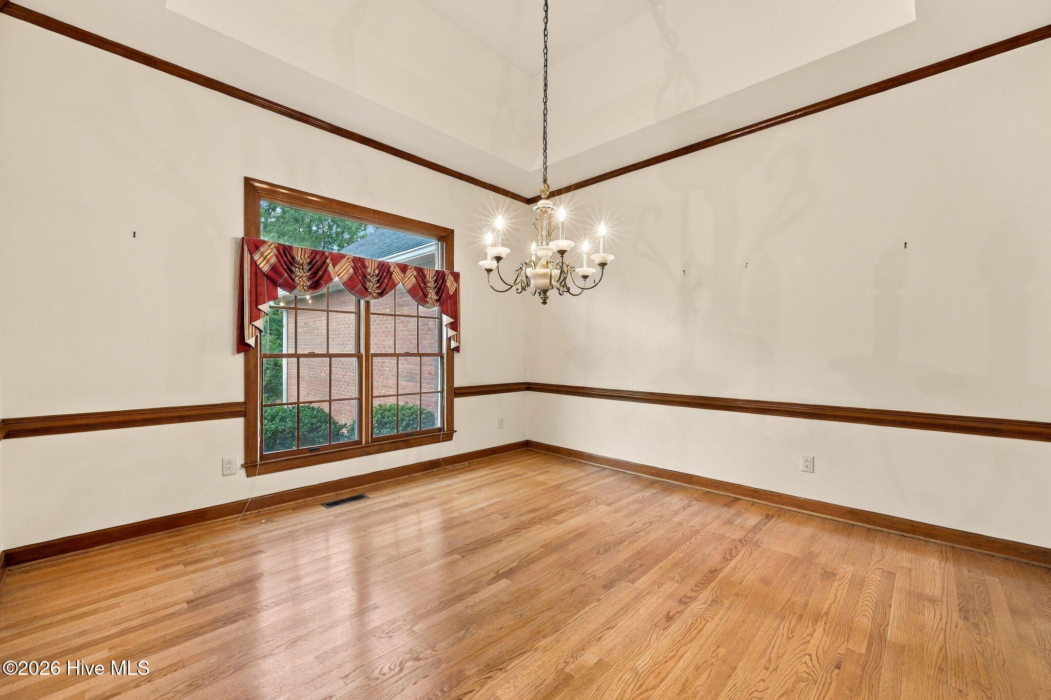 Empty room with hardwood floors, dark wood trim, a curtained window, and chandelier.