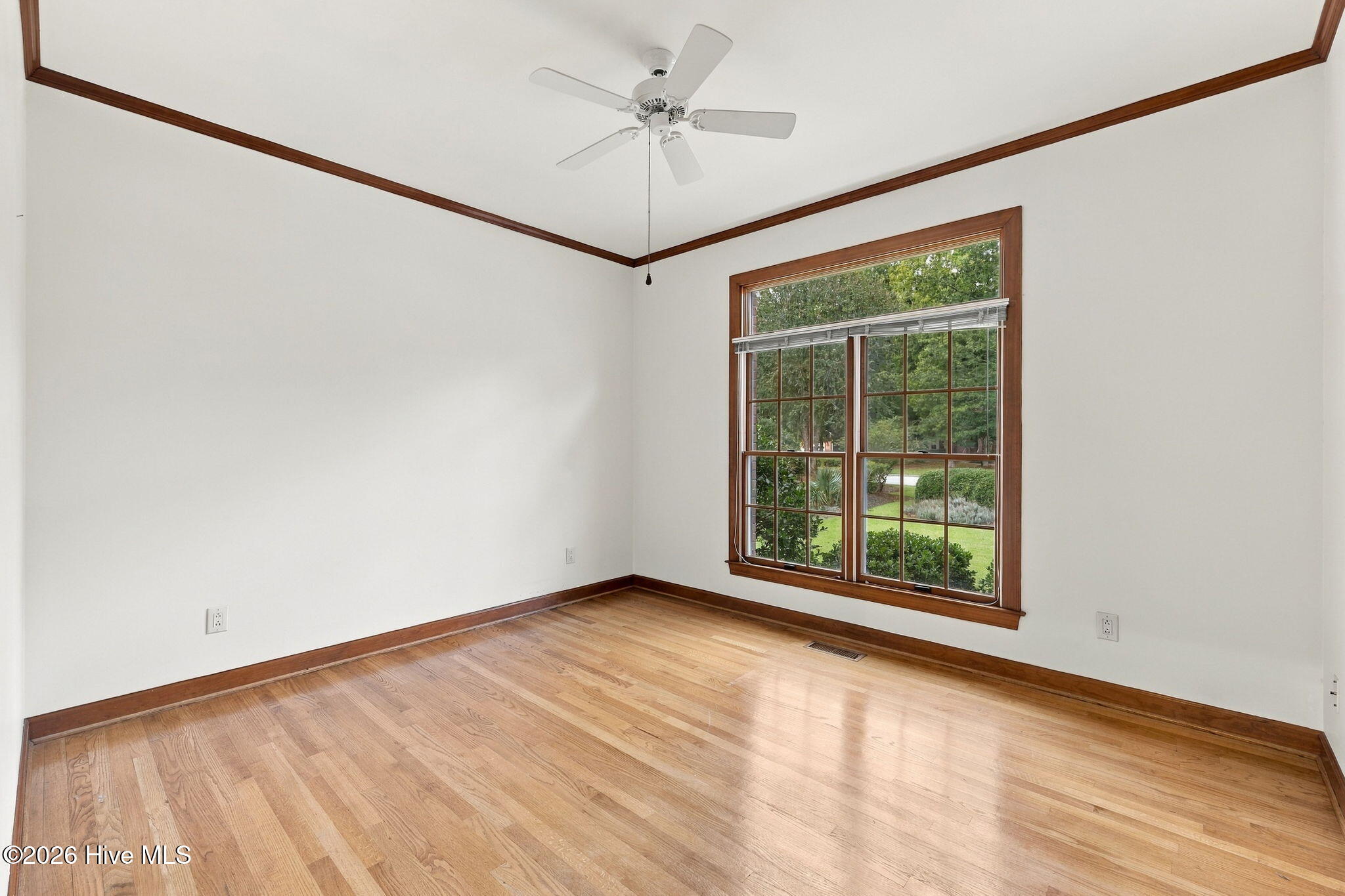 Empty room with hardwood floors, dark wood trim, ceiling fan, and a large window overlooking greenery.