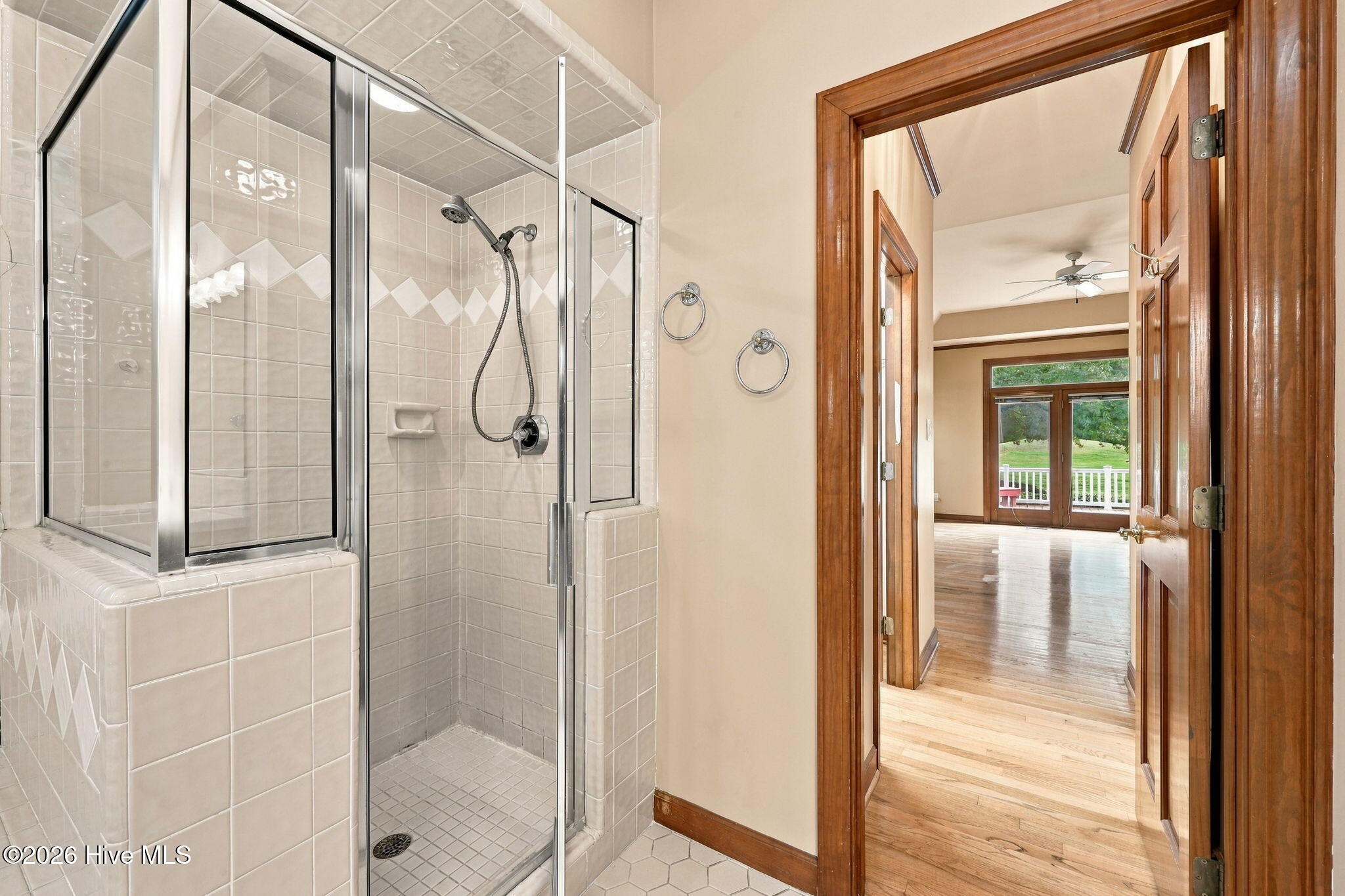 Tiled bathroom with glass shower and doorway overlooking a bright hardwood-floored room.
