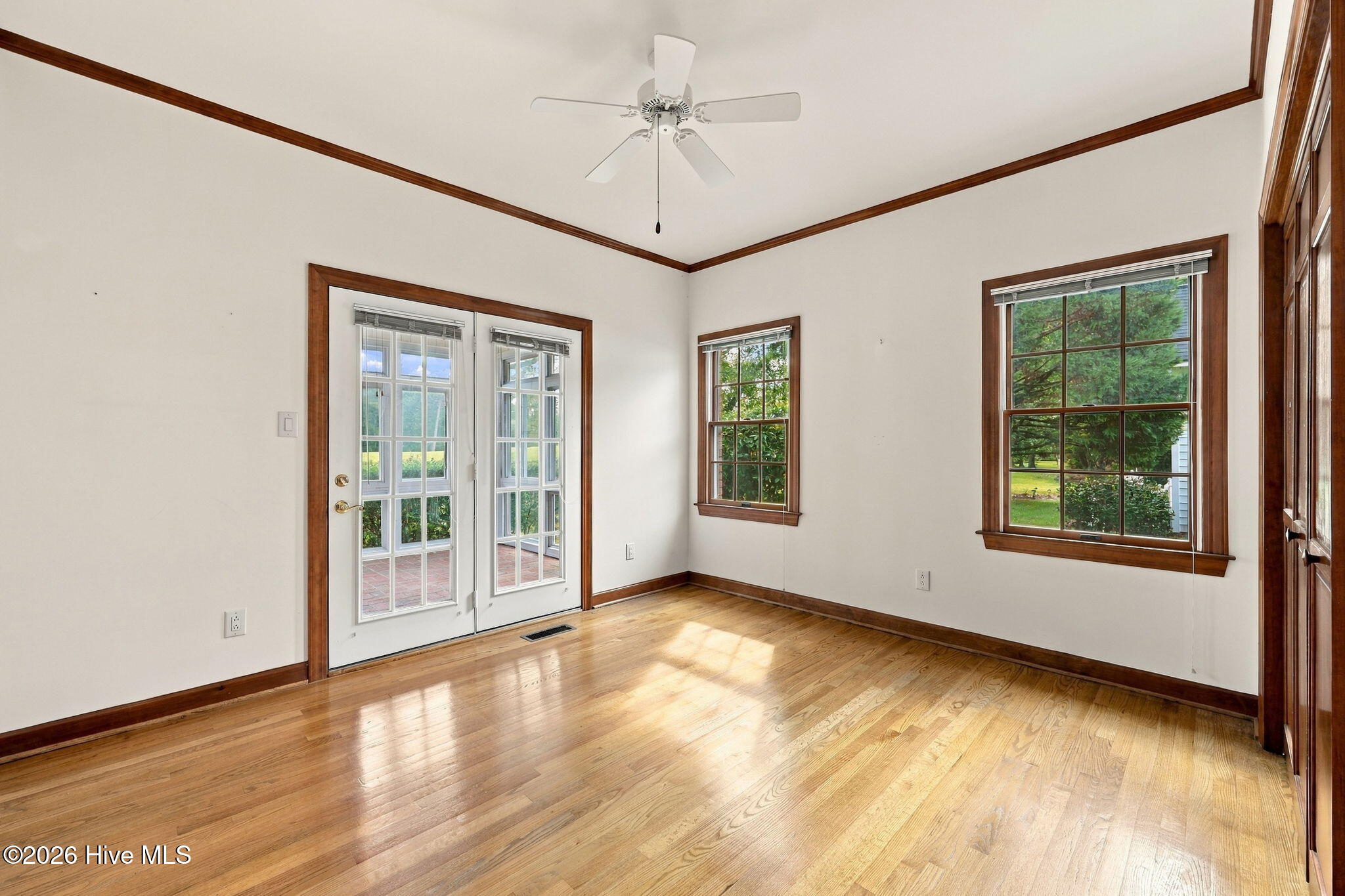 Empty sunlit room with hardwood floors, wood-trimmed windows, and glass doors opening toward a brick patio.