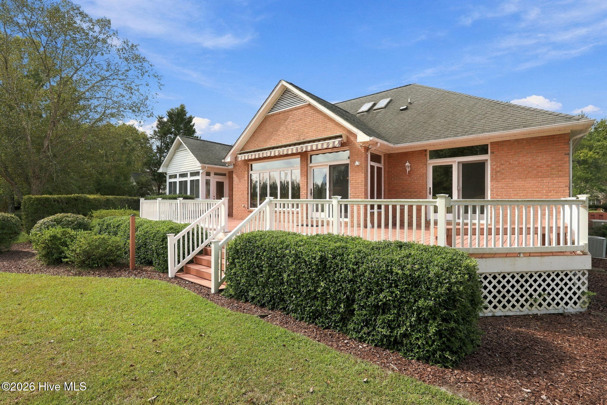 Brick home’s rear exterior with a raised white-railed deck, stairs, and landscaped shrubs.