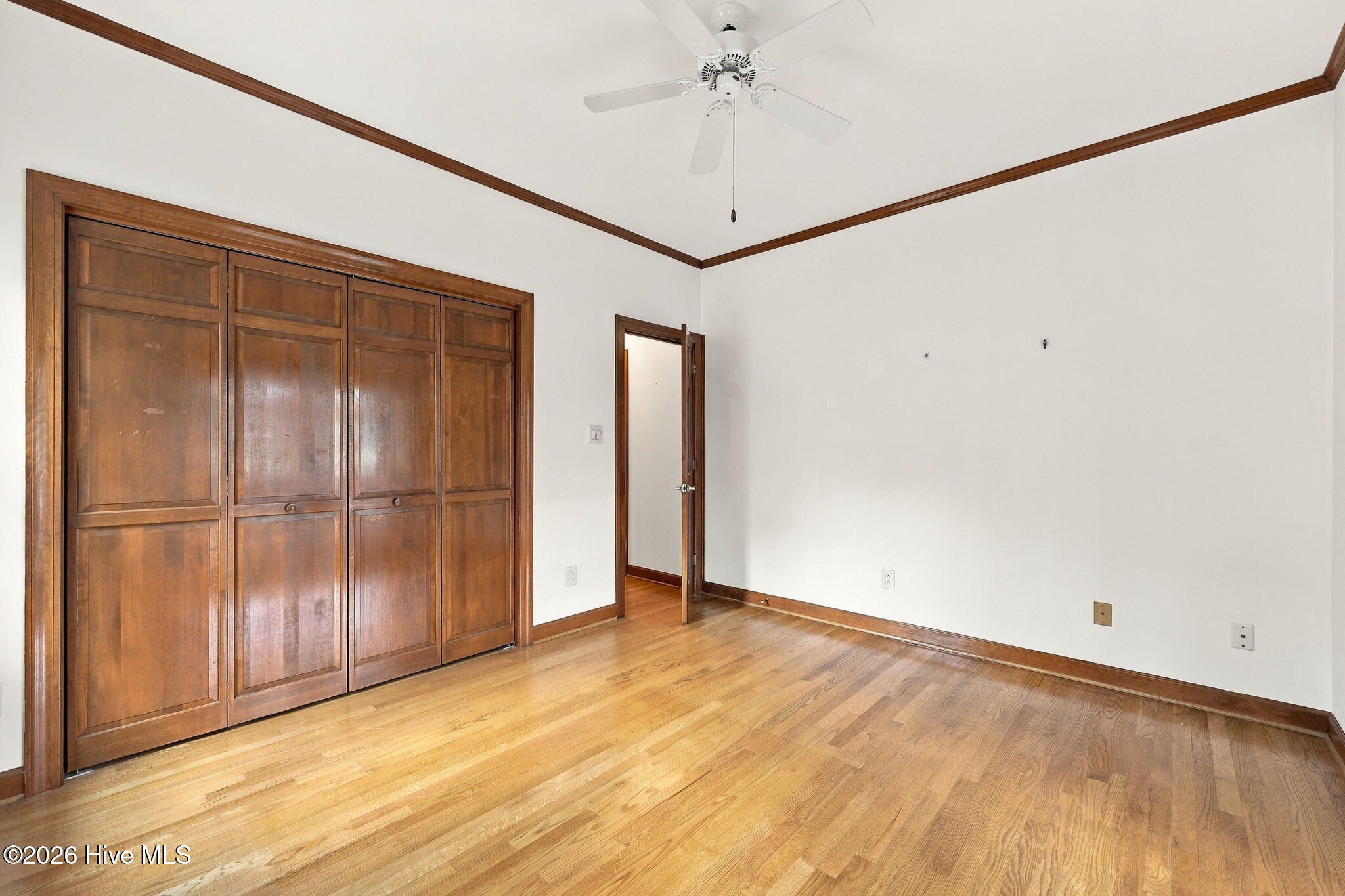 Empty bedroom with paneled wooden closets, hardwood floors, and a ceiling fan.