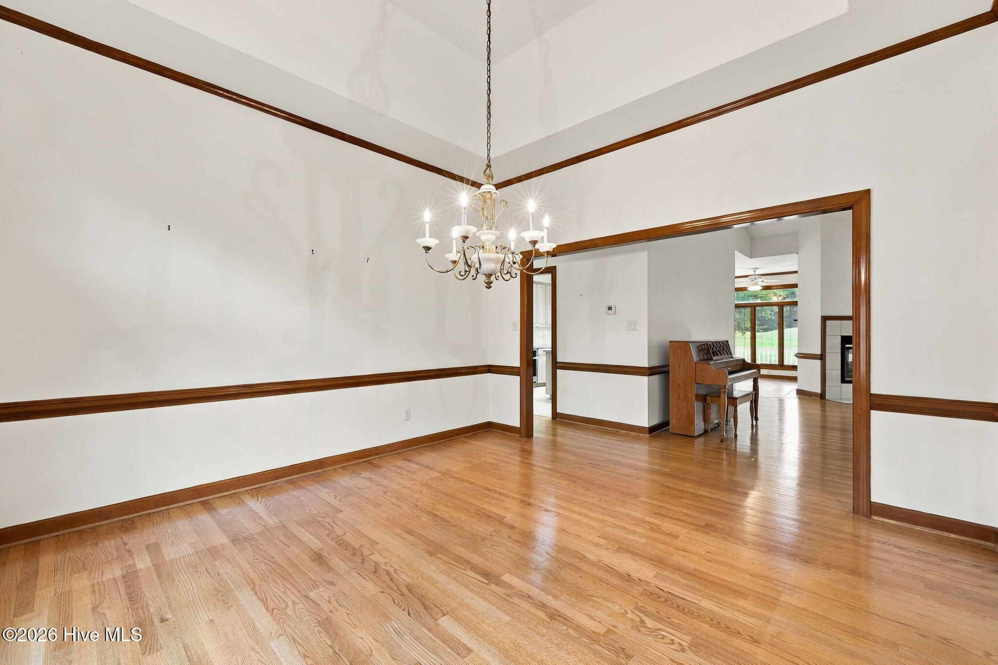 Dining room with hardwood floors and chandelier, opening to a living area with a piano and fireplace.