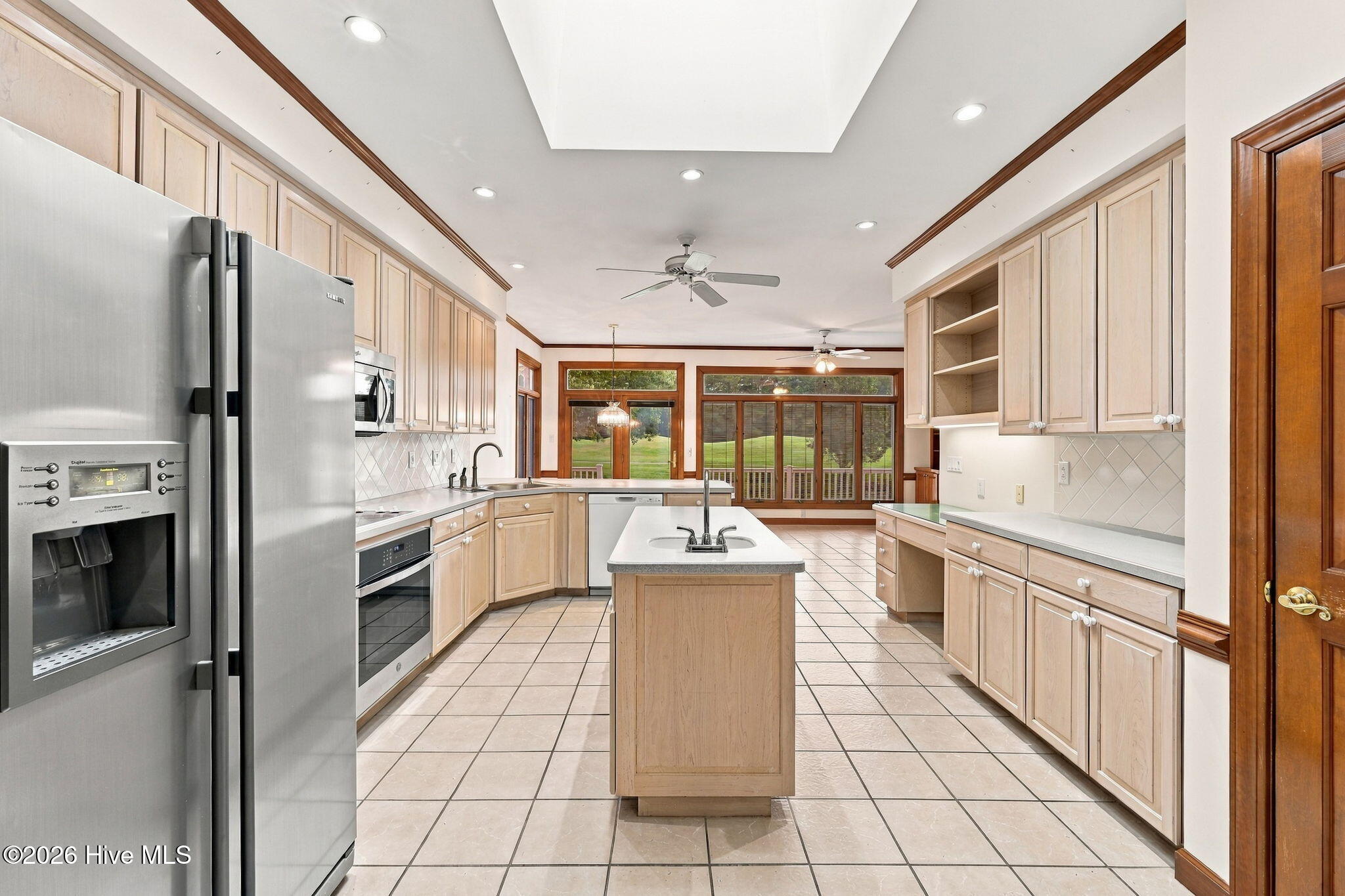 Spacious kitchen with a central sink island, light wood cabinets, stainless steel appliances, and windows overlooking a lawn.