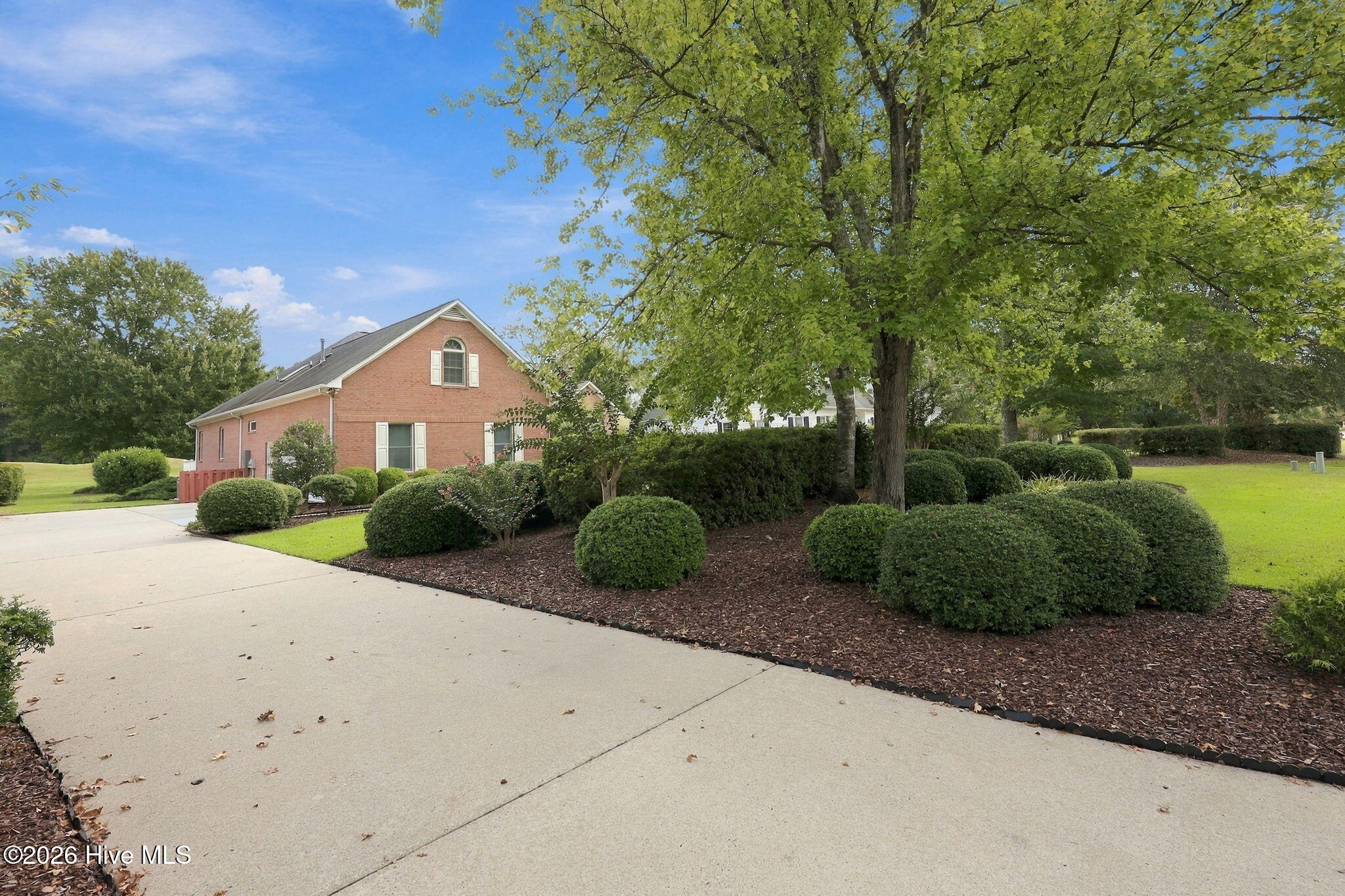 Red-brick home beyond a wide driveway, framed by mature trees and neatly trimmed shrubs.