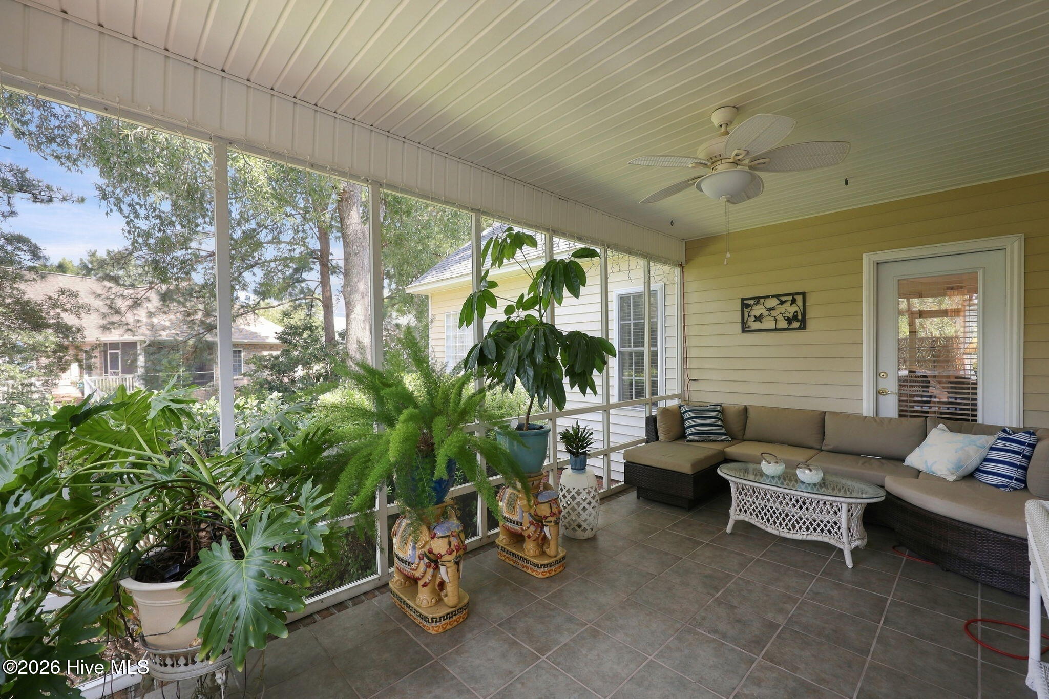 Screened porch with tiled floors, a beige sectional, ceiling fan, and abundant potted plants.