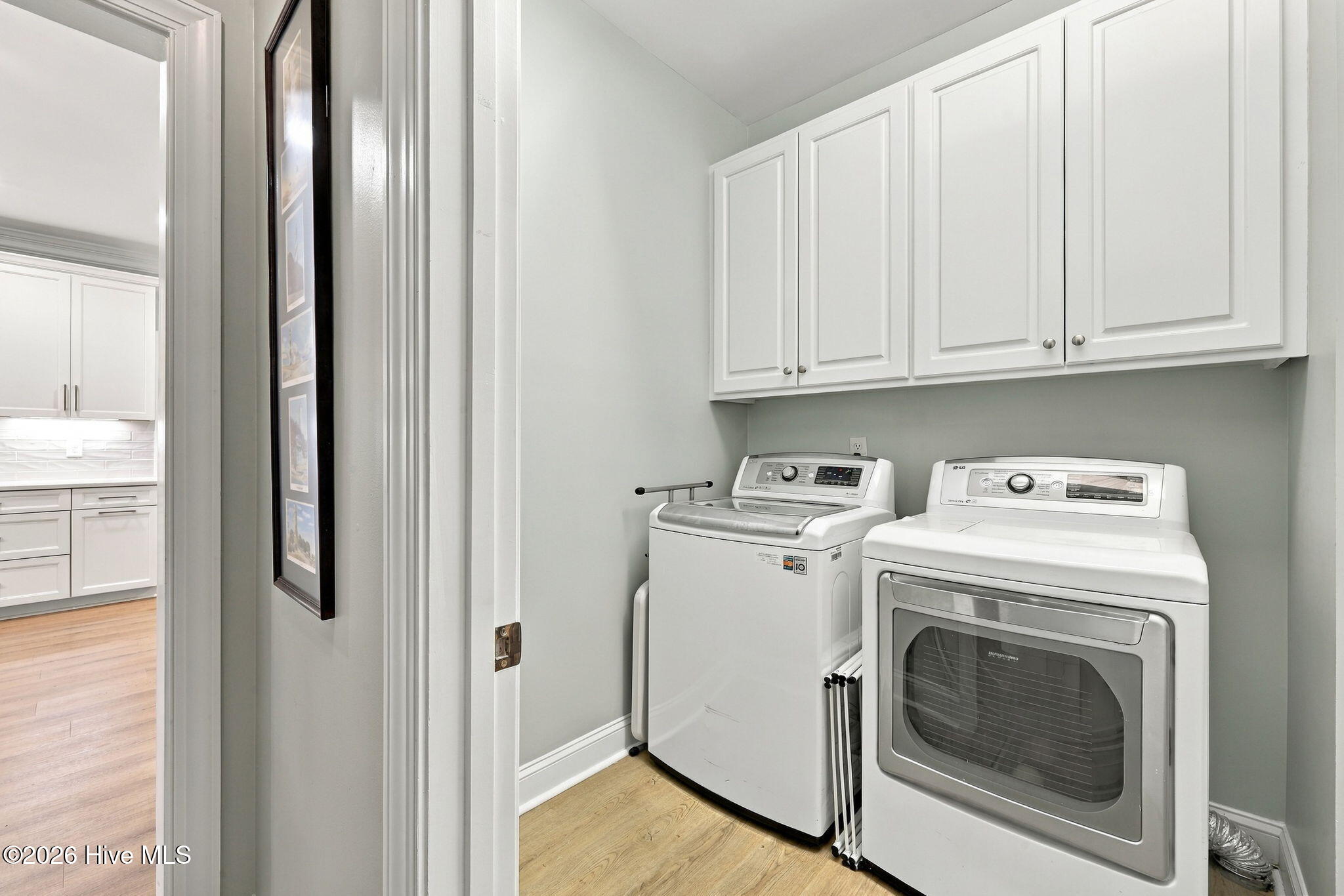 Laundry room with white washer and dryer beneath wall cabinets, next to a kitchen doorway.