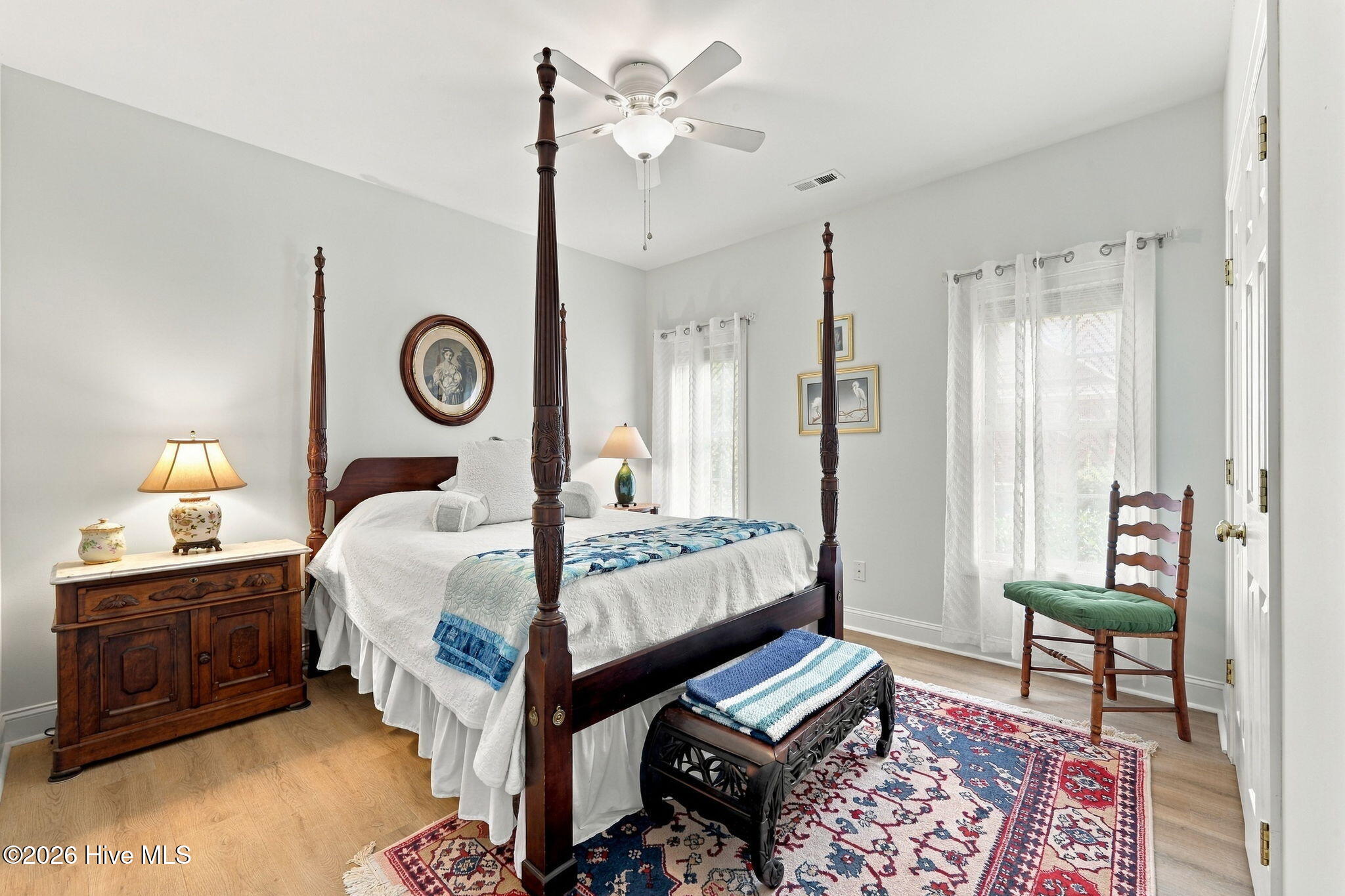 Furnished bedroom with a carved four-poster bed, sheer-curtained windows, and a colorful patterned rug.