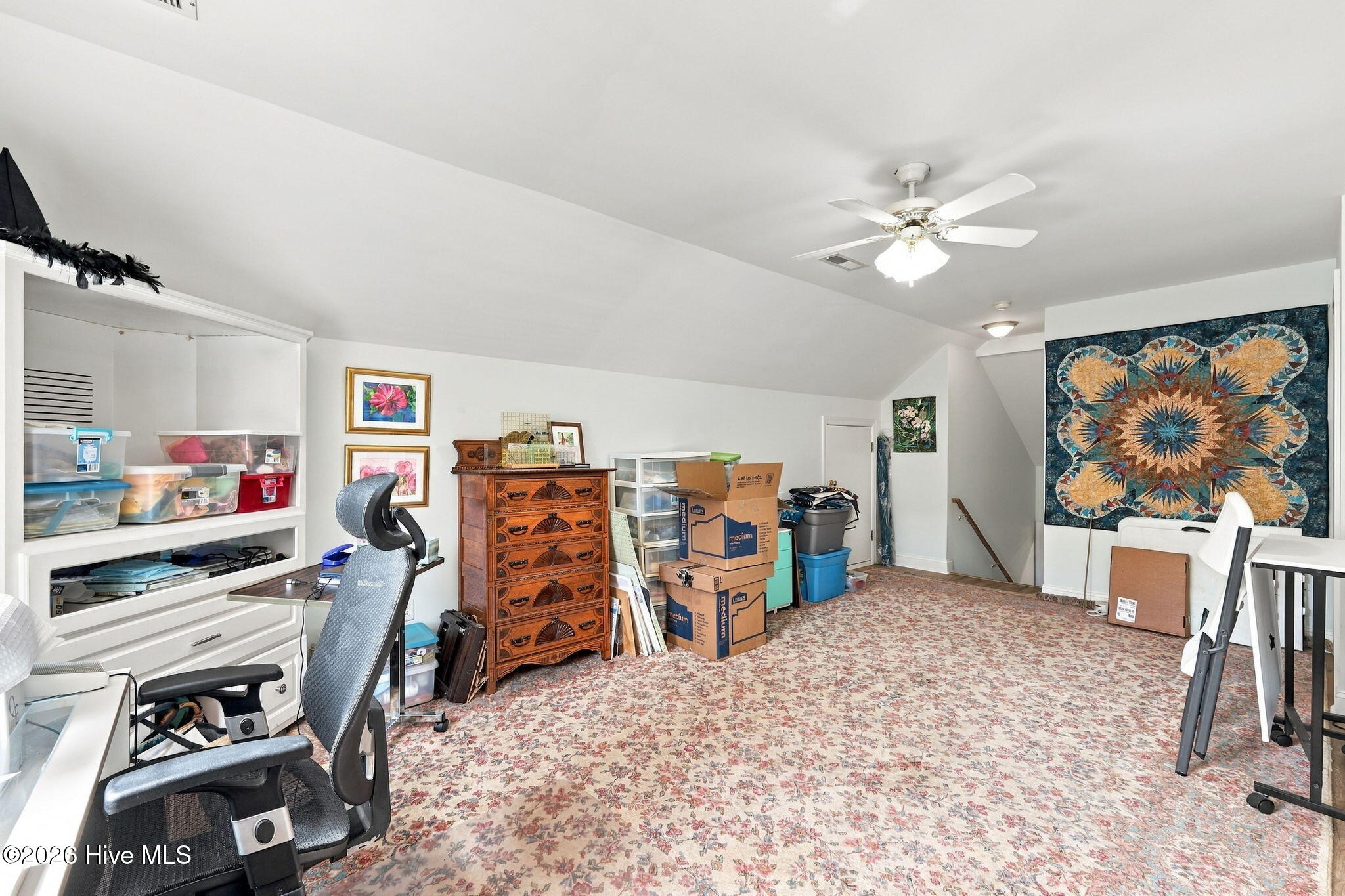 Carpeted bonus room with built-in storage, moving boxes, an office chair, and a colorful quilt on the wall.