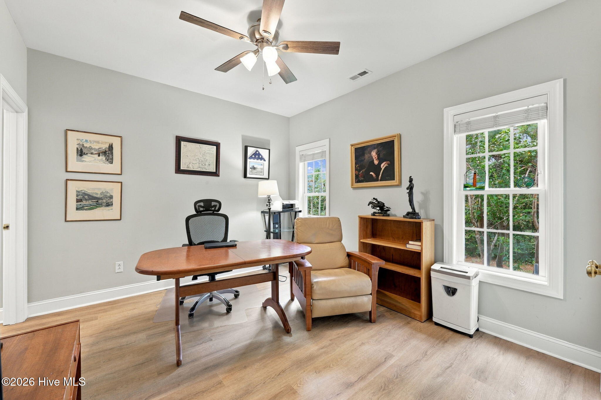 Home office with a wooden desk, tan armchair, framed artwork, and windows overlooking greenery.