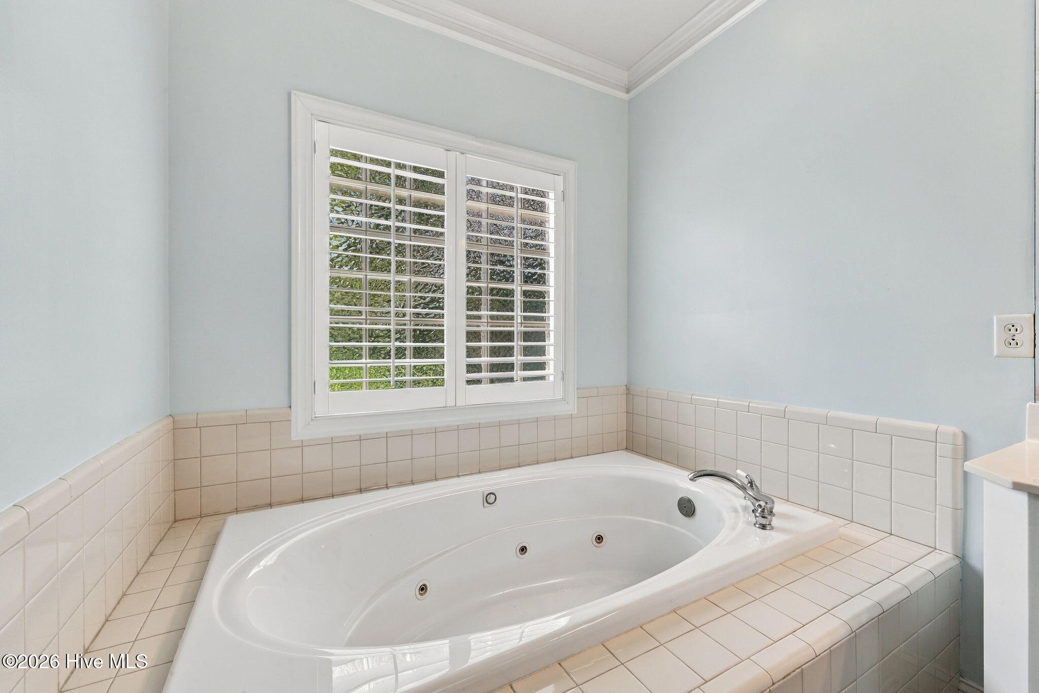 Jetted soaking tub beneath plantation-shuttered window in a light blue bathroom.