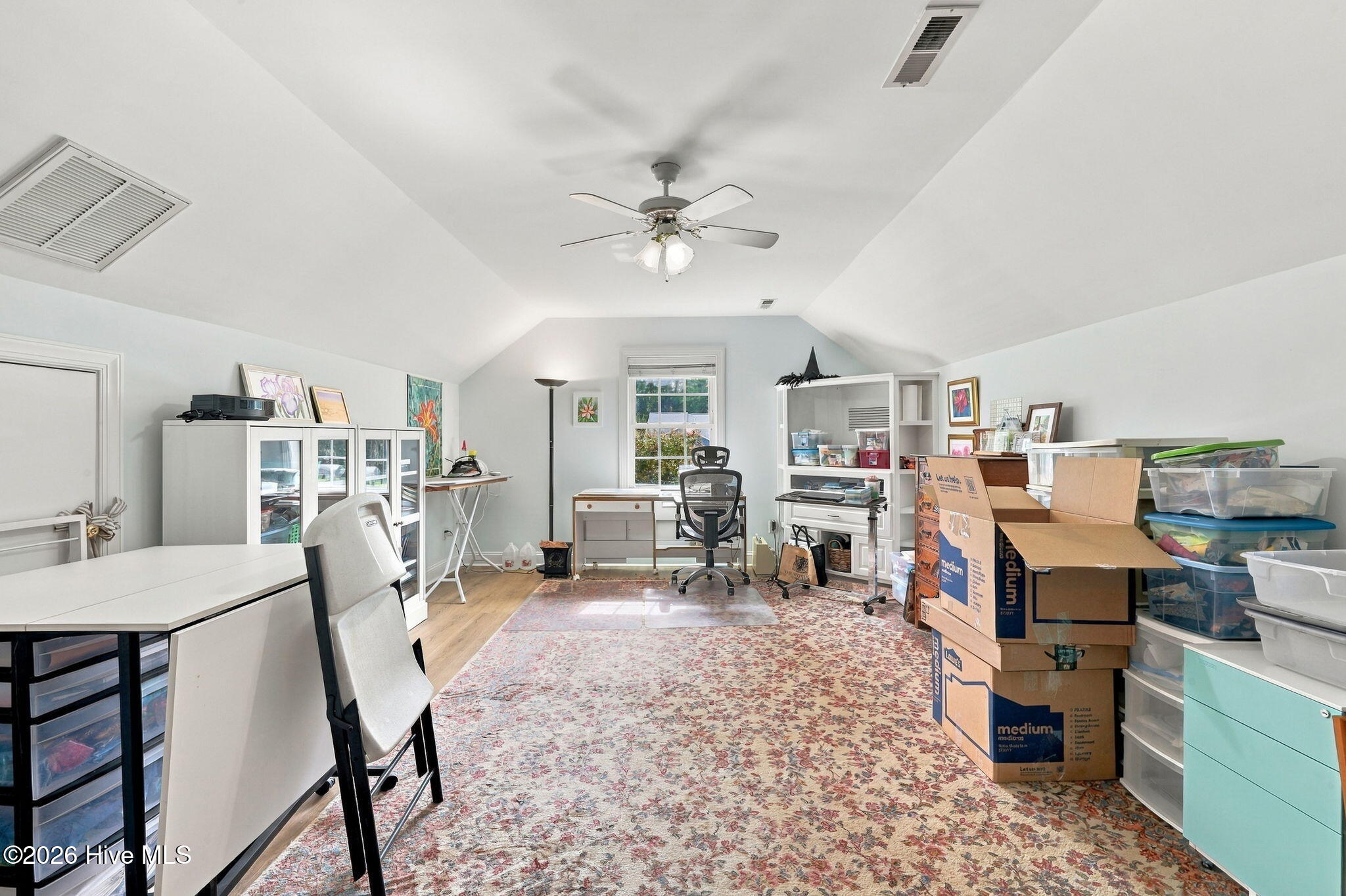 Home office with a vaulted ceiling, desk, patterned rug, and stacked moving boxes.
