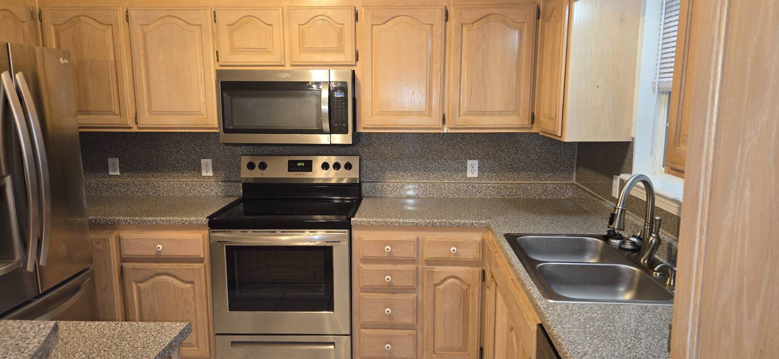 Kitchen with light wood cabinets, speckled counters, stainless steel appliances, and a double-basin sink.