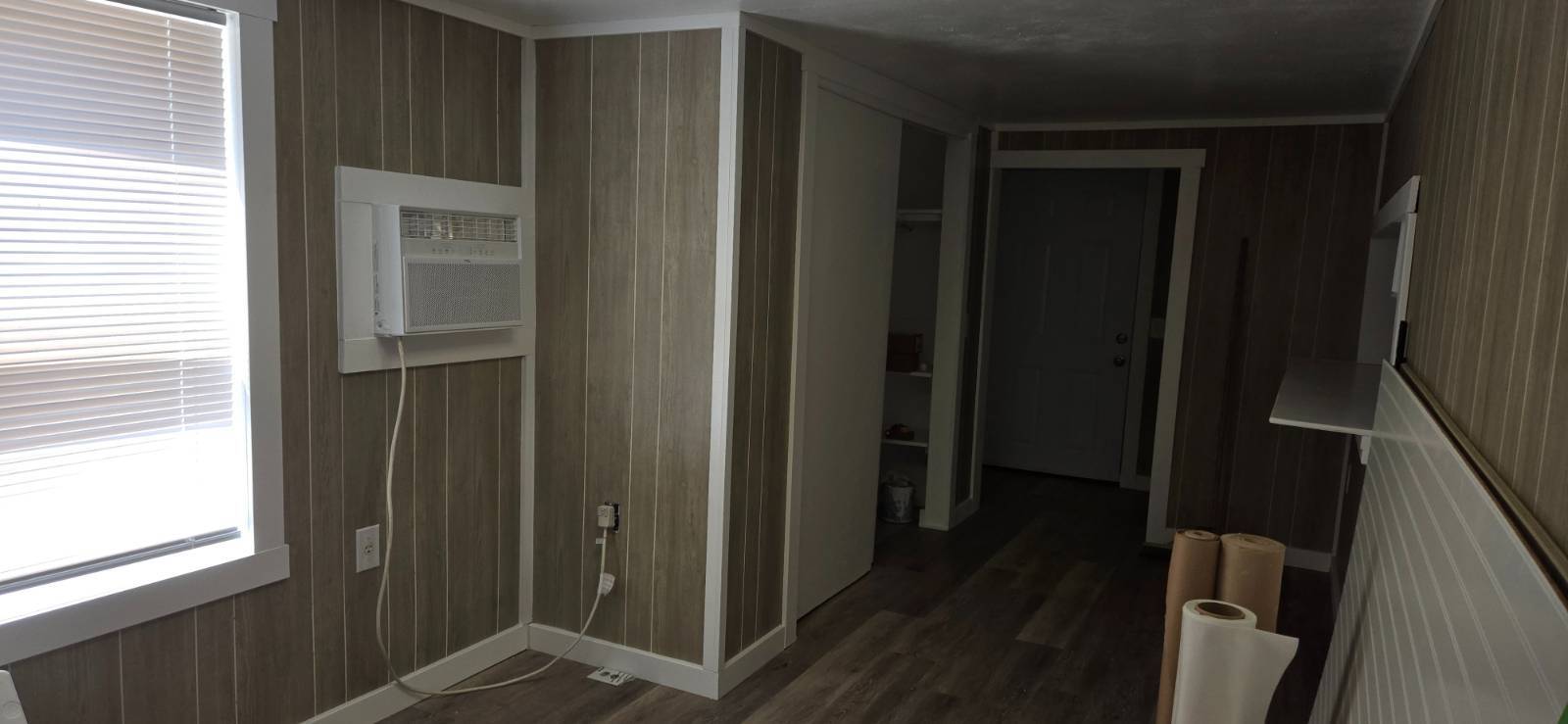 Empty paneled room with a window air conditioner, wood-look flooring, and an open hallway.