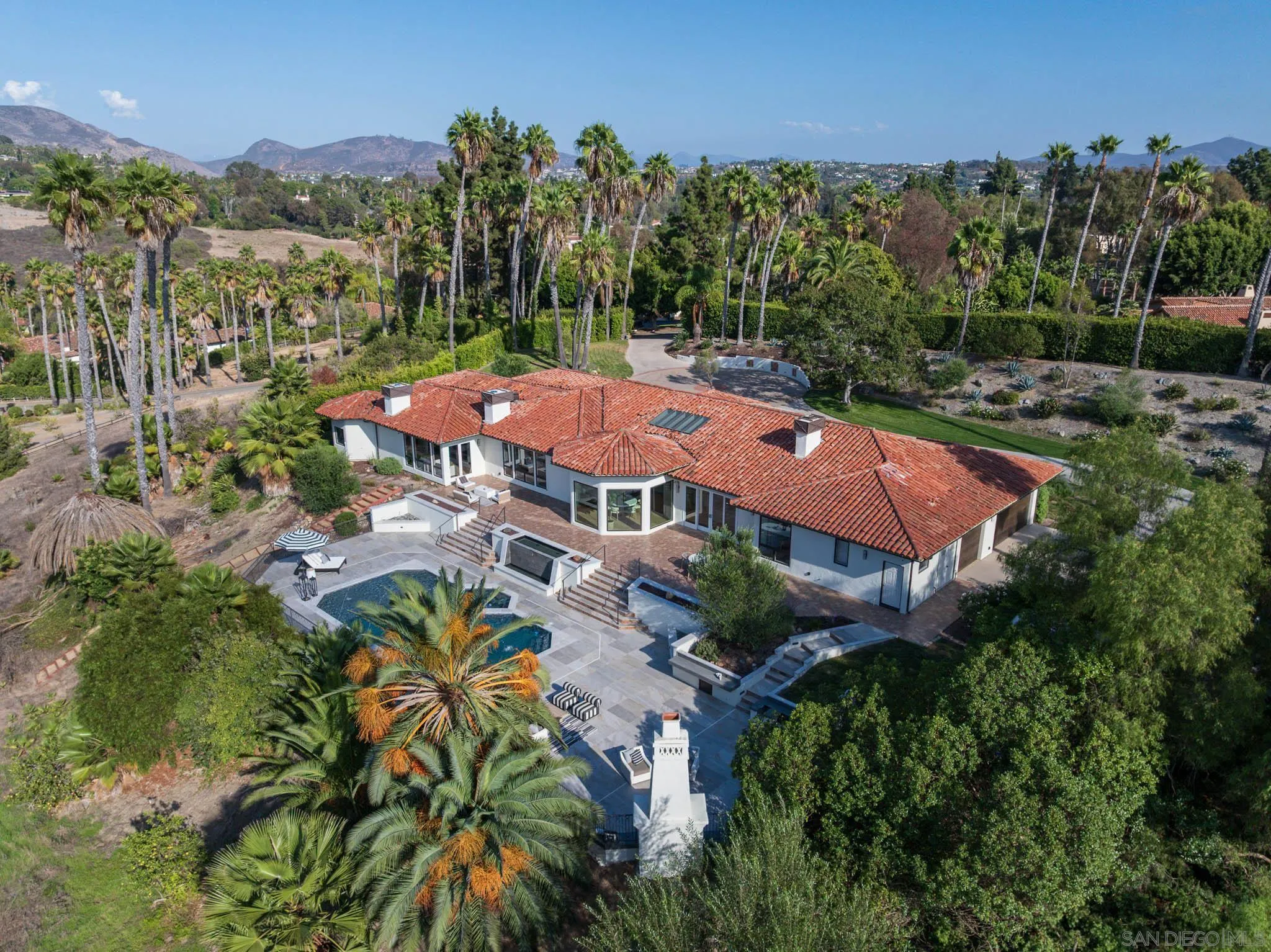 Aerial view of a tile-roofed home with a swimming pool, palm trees, and mountain backdrop.