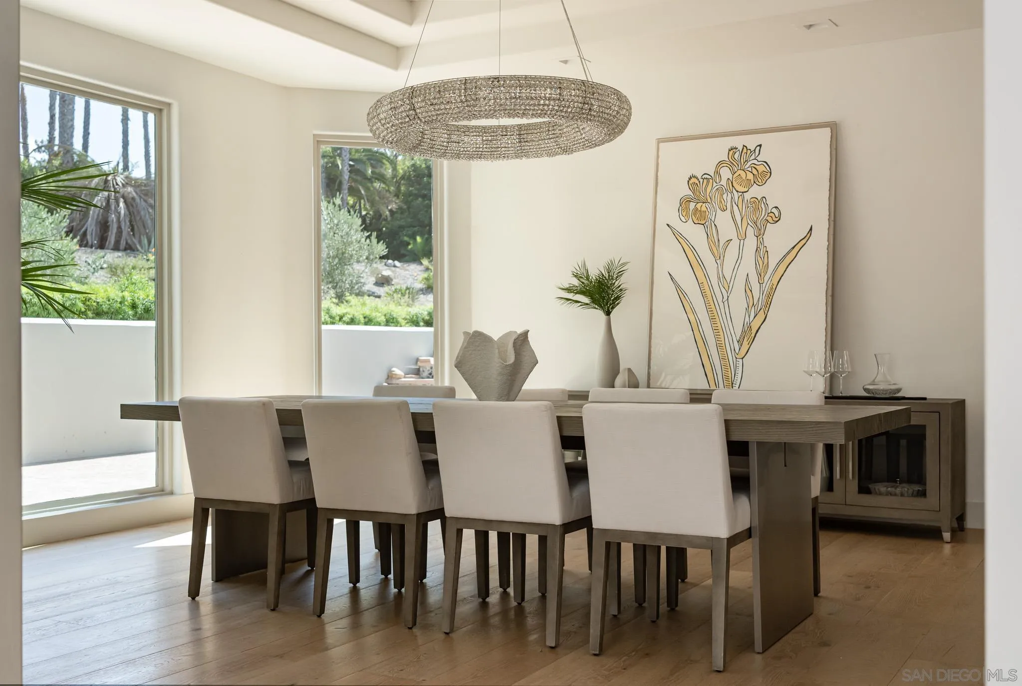 Dining room with a long wood table, upholstered chairs, crystal chandelier, and tall garden-facing windows.