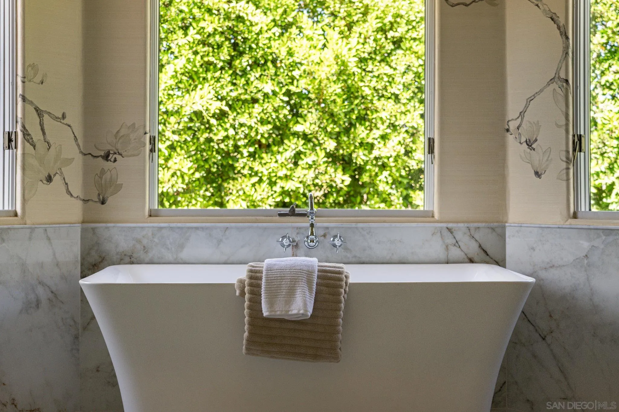 Freestanding bathtub with marble surround, towels, and a garden view through the window.